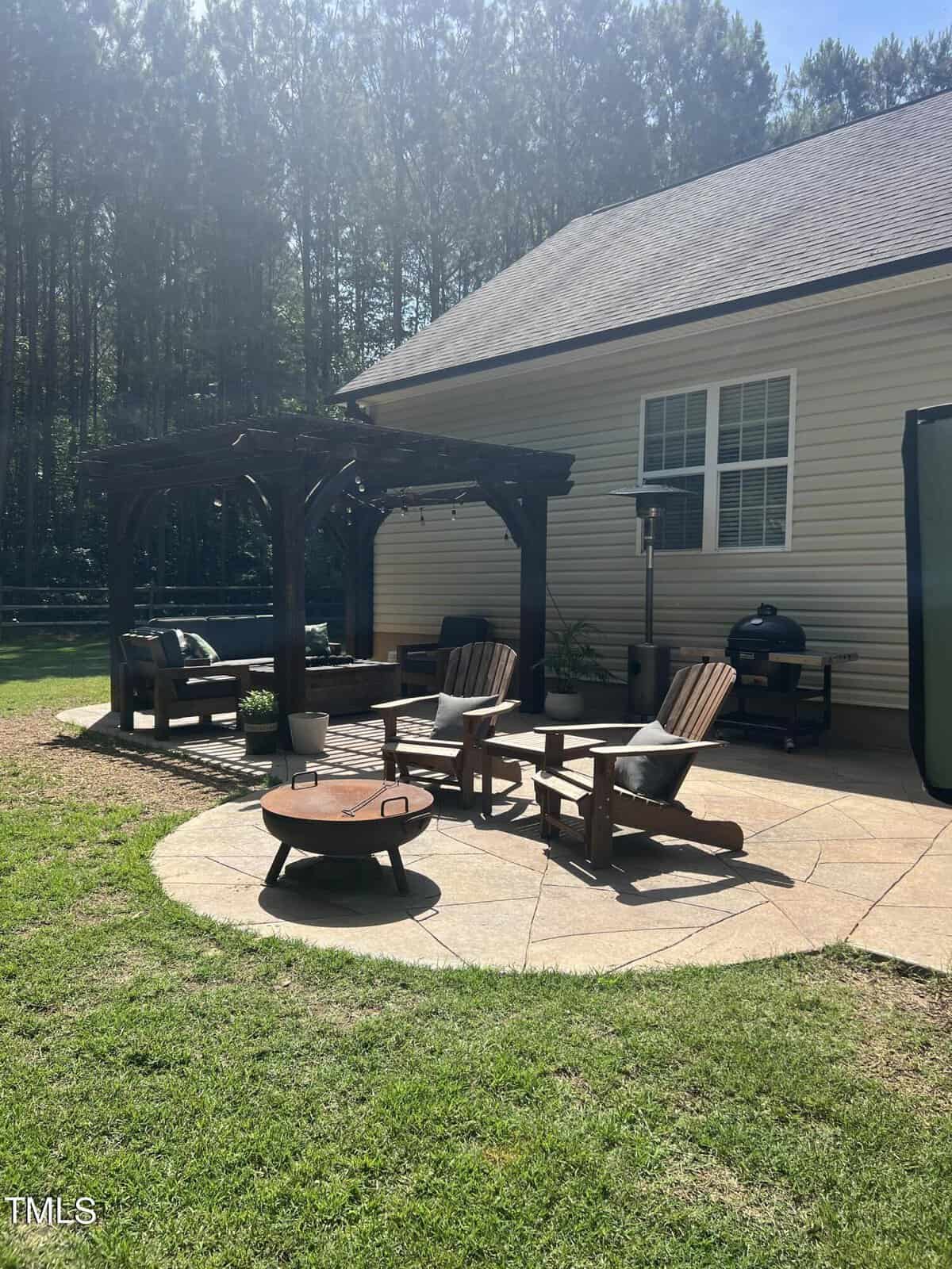 A backyard patio with wooden Adirondack chairs, a fire pit, and a covered pergola. A grill and patio heater are near the house, and trees line the background, creating a serene outdoor space.