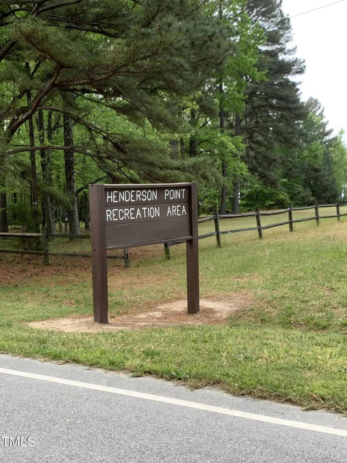 A wooden sign reading "Henderson Point Recreation Area" stands beside a road with a wooden fence and tall, green trees in the background. The ground around the sign is grassy and partially bare of vegetation.