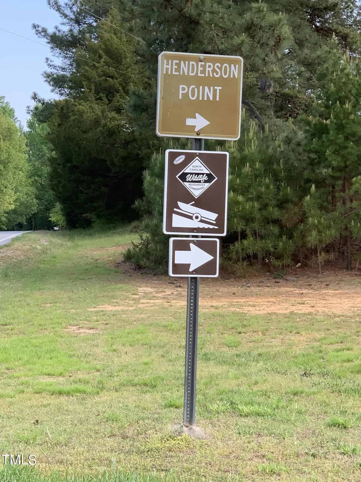 A signpost stands on a grassy area beside a road, indicating "Henderson Point" in bold white letters on a brown sign. Below it, there are additional directional signs with arrows pointing to the right, leading the way. Trees and a road can be seen in the background.