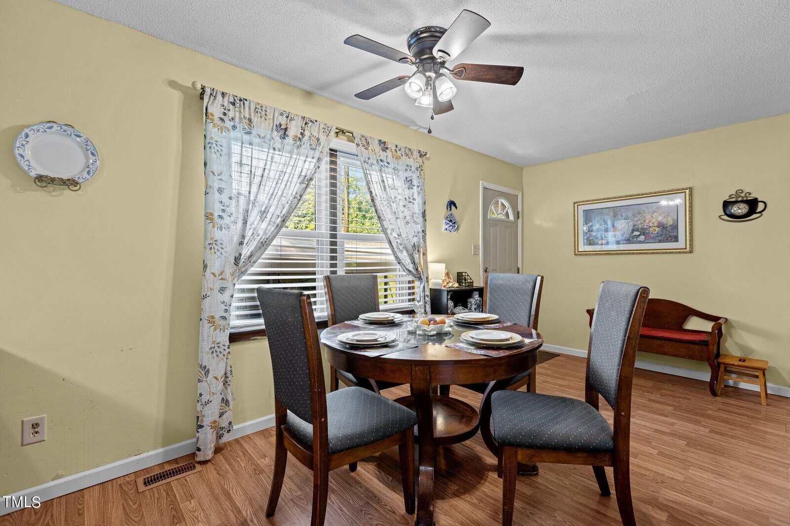 A dining room with a round wooden table set for four people. The table has grey upholstered chairs, plates, and a small centerpiece. There is a window with floral curtains, a ceiling fan, a decorative plate on the wall, artwork, and a wooden bench in the background.