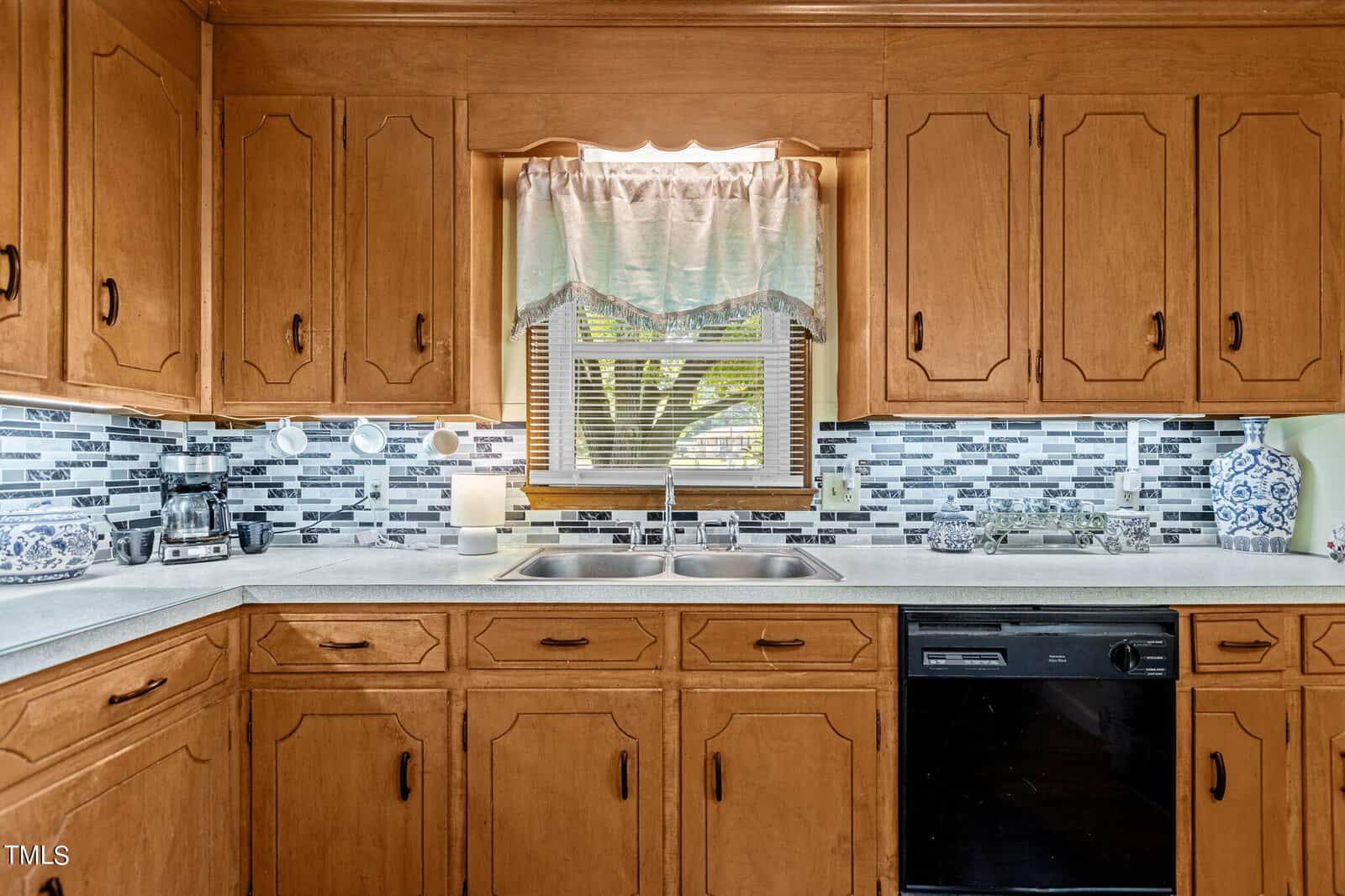 A kitchen featuring wooden cabinets, a tiled backsplash in shades of grey and white, and a stainless steel double sink under a window with a lace curtain. Appliances, including a dishwasher, coffee maker, and microwave, are situated on the countertop.