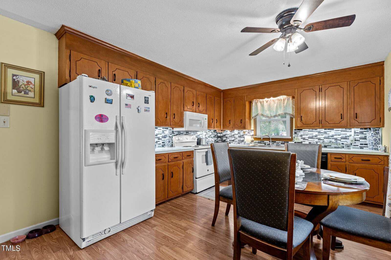 A cozy kitchen featuring wooden cabinets, a white refrigerator with magnets, and a white stove. A ceiling fan with light illuminates the space. A wooden dining table with four black chairs is set with plates, and a mosaic tile backsplash adds a modern touch.
