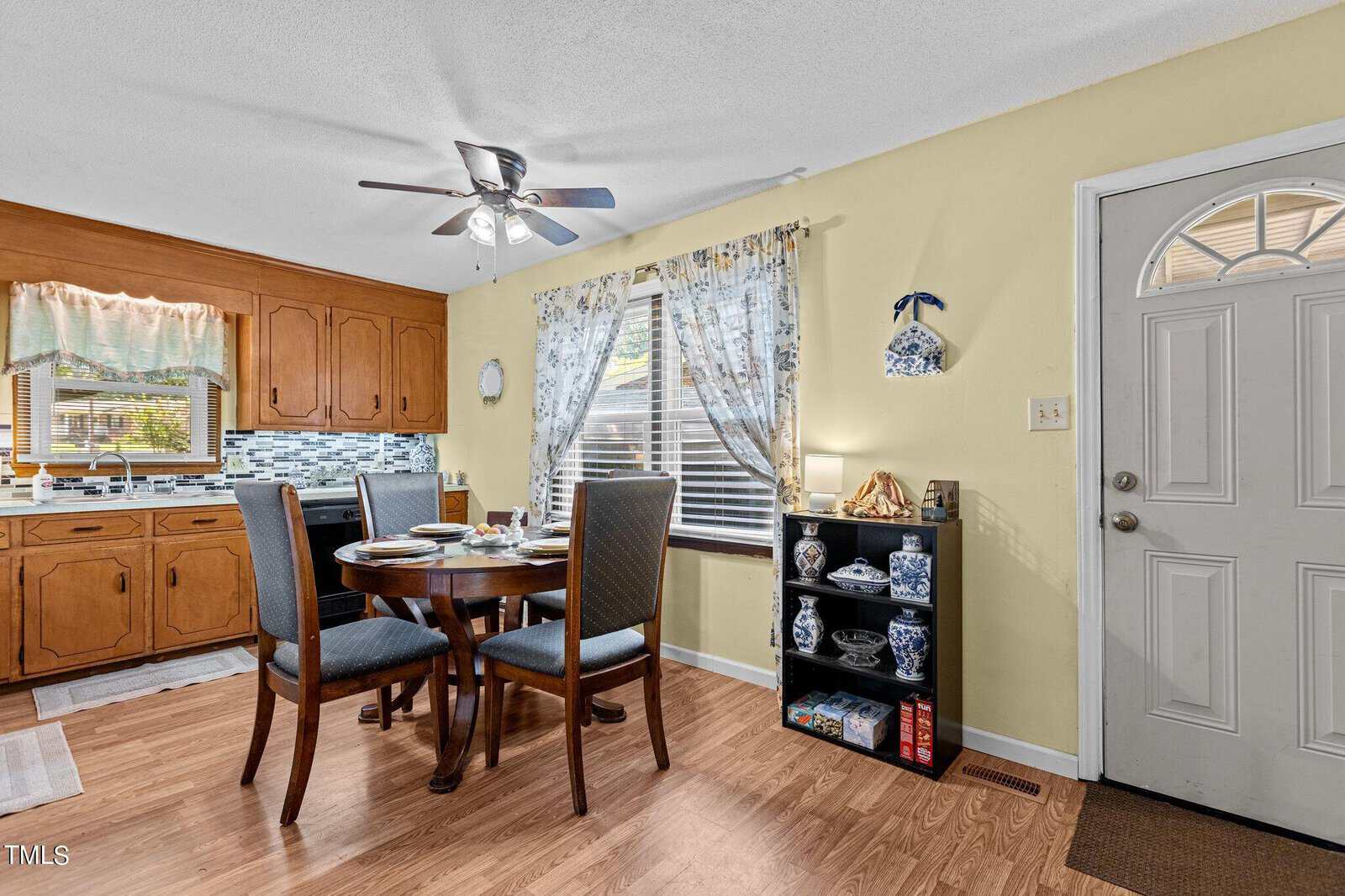 A cozy kitchen with wooden cabinets and a backsplash of blue and white tiles. A round wooden dining table with four chairs is set against a window with floral curtains. To the right, there's a shelf with decorative plates and a white door with a window at the top.