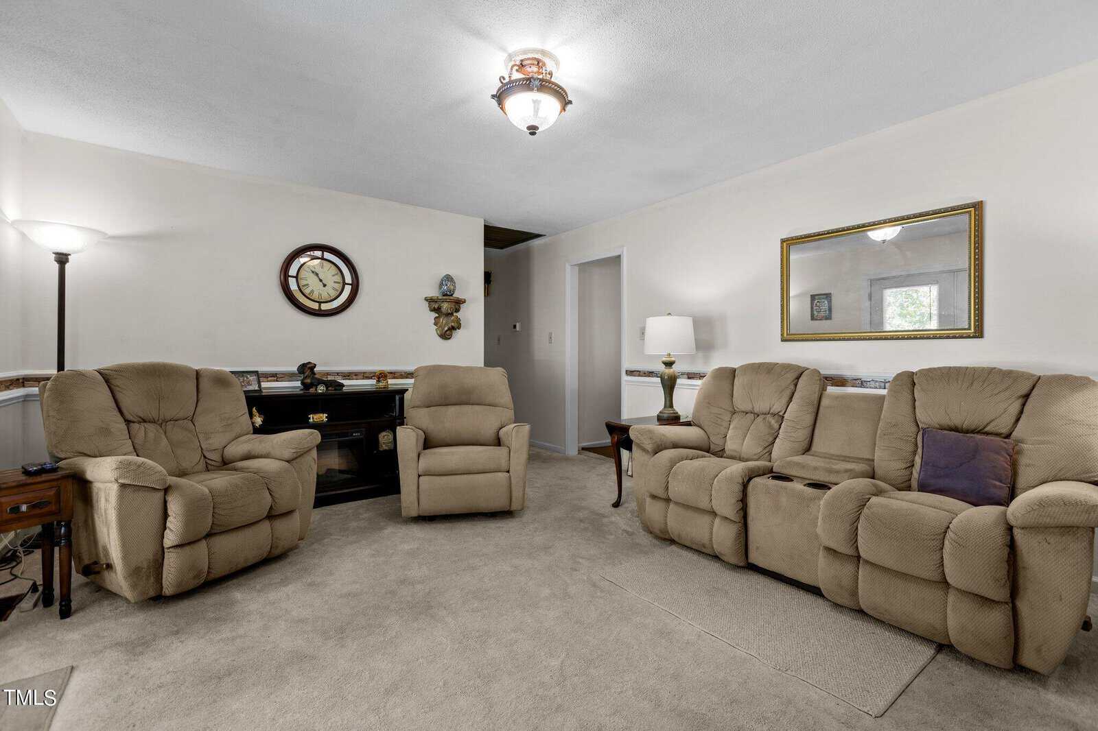 A cozy living room with beige carpet features three plush, light brown recliners. There's a dark wooden desk with a clock and wall decor above it. A large mirror hangs on the pale wall, and soft lighting comes from a ceiling fixture and floor lamp.