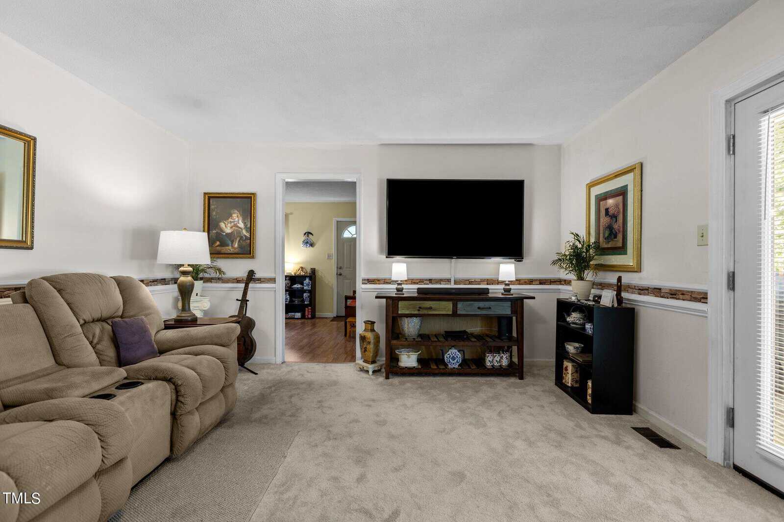 A cozy living room with a beige recliner sofa, a TV mounted on the wall above a wooden console with shelves, and a lamp on one side of the console. A door on the left leads to another room. The room is decorated with framed pictures and has a light-colored carpet.