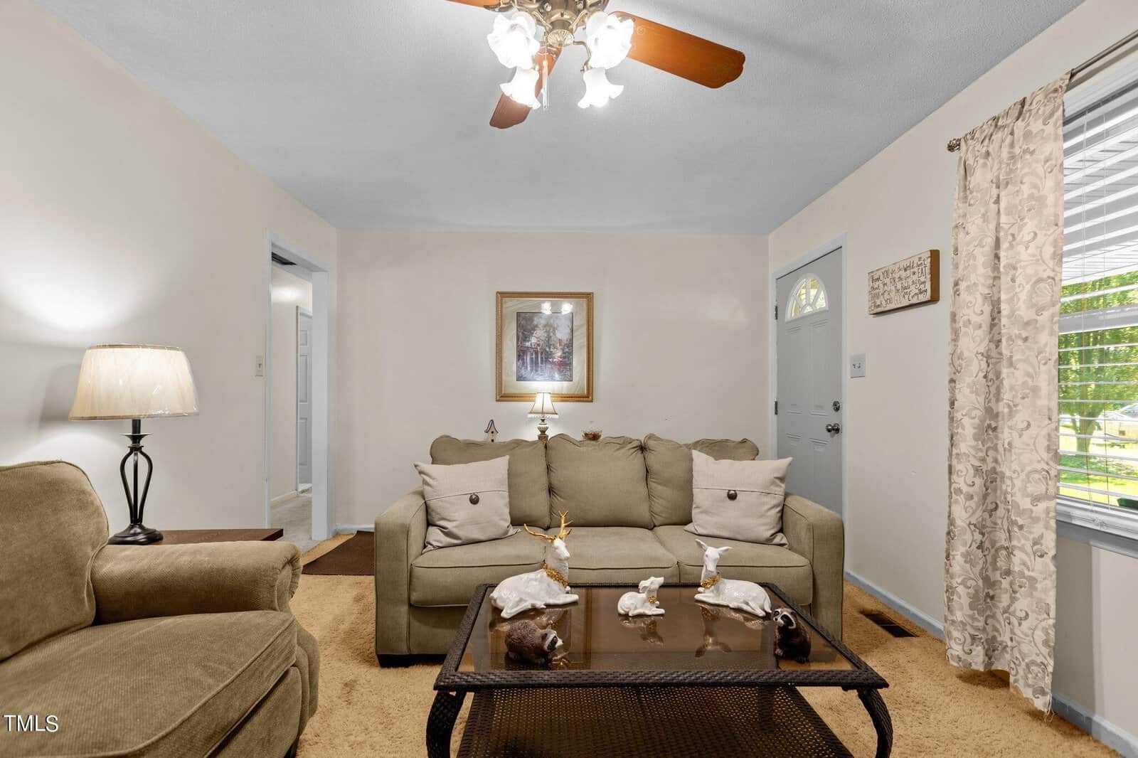 A cozy living room with beige walls and a ceiling fan with four wooden blades and light fixtures. The room features a beige sofa, a matching armchair, a glass coffee table, and a lamp. A framed picture hangs on the wall above the sofa, and a window with curtains is on the right.