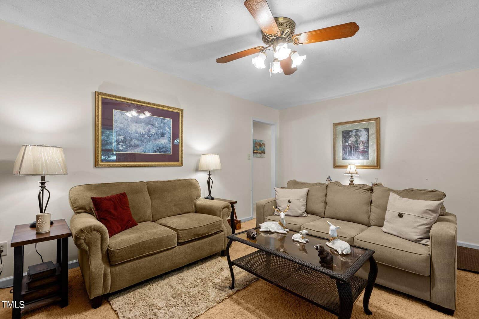 A cozy living room with a brown sofa and loveseat, adorned with beige and red pillows. Two lamps on side tables flank the sofa. A coffee table displays various decorations. Framed artwork hangs on light-colored walls, and a ceiling fan with lights is overhead.