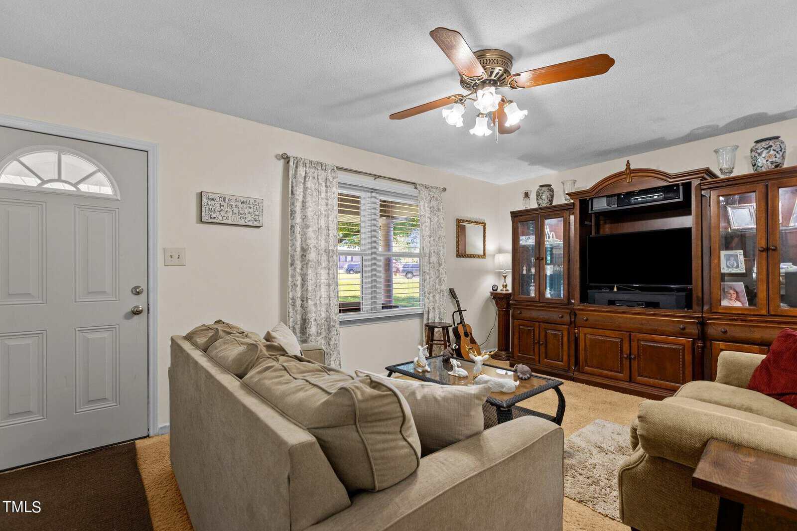 A cozy living room with a beige couch and a coffee table topped with books and decor. A wooden entertainment unit with a TV and shelves holding various items stands against the wall. A ceiling fan and large window bring light into the space. A guitar leans near the window.