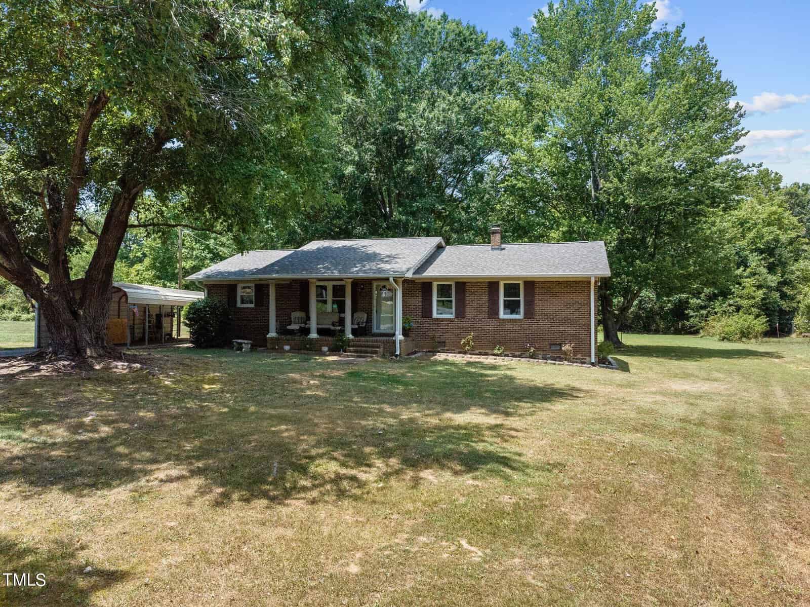 A single-story brick house with a gable roof is set amidst lush greenery. It features a covered front porch supported by columns and a carport on the left. The yard is expansive, with a large tree providing shade near the house and a well-kept lawn extending outward.