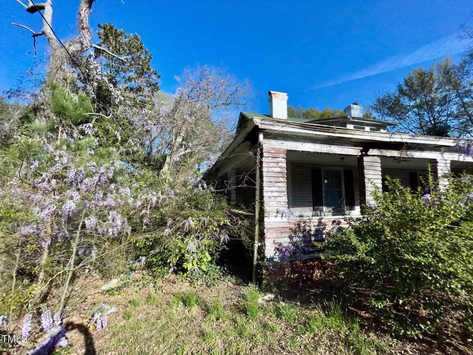 An old, abandoned wooden house at 925 Nicholas stands amidst overgrown vegetation and purple wisteria vines in Henderson, NC. The roof is slightly damaged, and the walls are weathered. A bright blue sky with light clouds stretches out above the scenery.