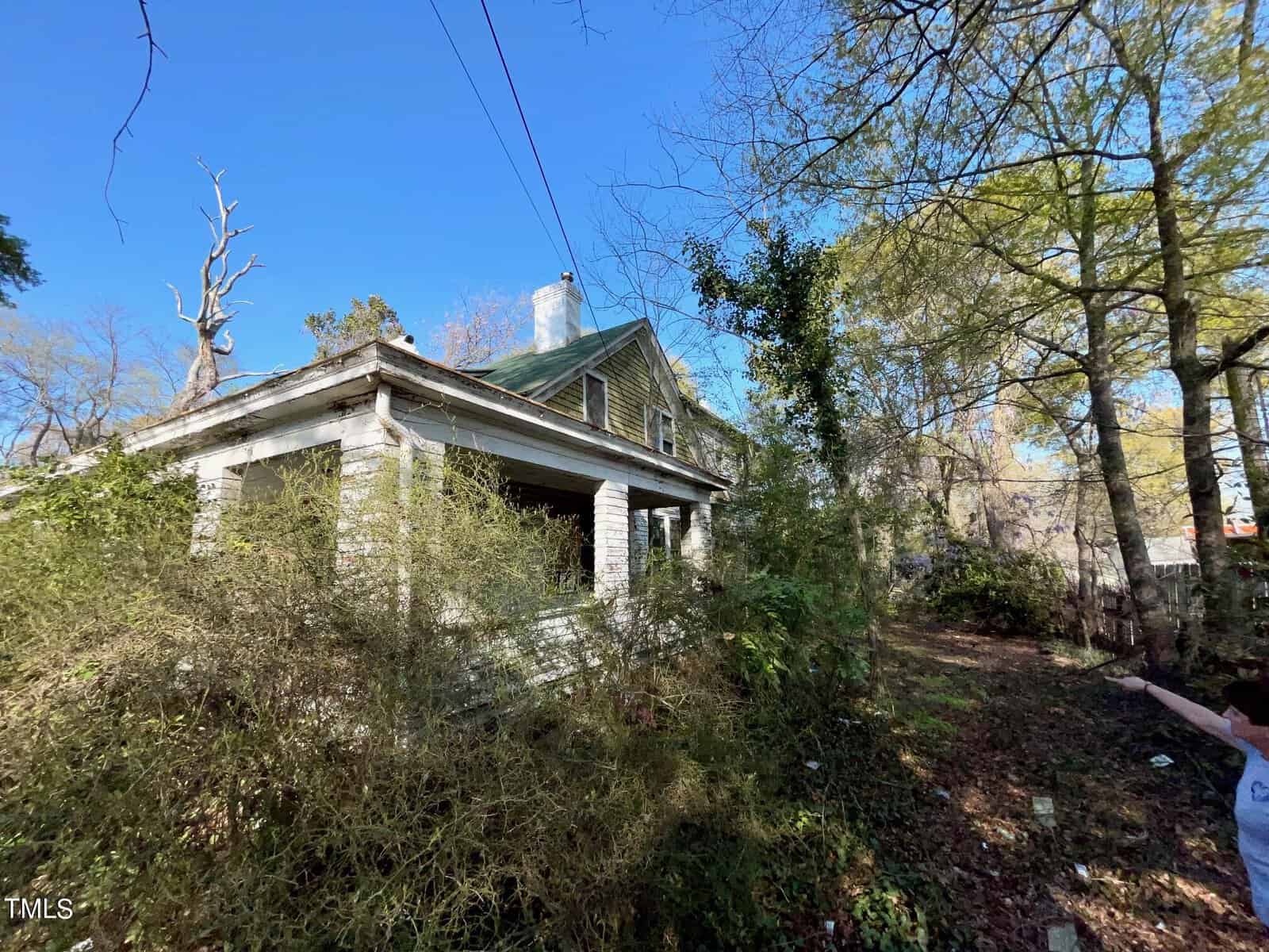 An abandoned, dilapidated house is partially hidden by overgrown plants and trees. The structure shows significant wear and tear, with peeling paint and broken windows. A person points towards the house from the right side of the image. The sky is clear and blue.