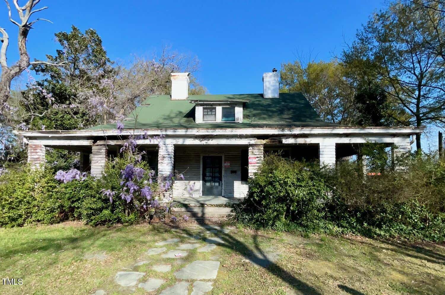 An old, rustic single-story house with a green roof and a large front porch. The exterior is weathered, and it is surrounded by overgrown vegetation, including bushes and tall trees. There is a stone pathway leading to the porch steps. The sky is clear and blue.