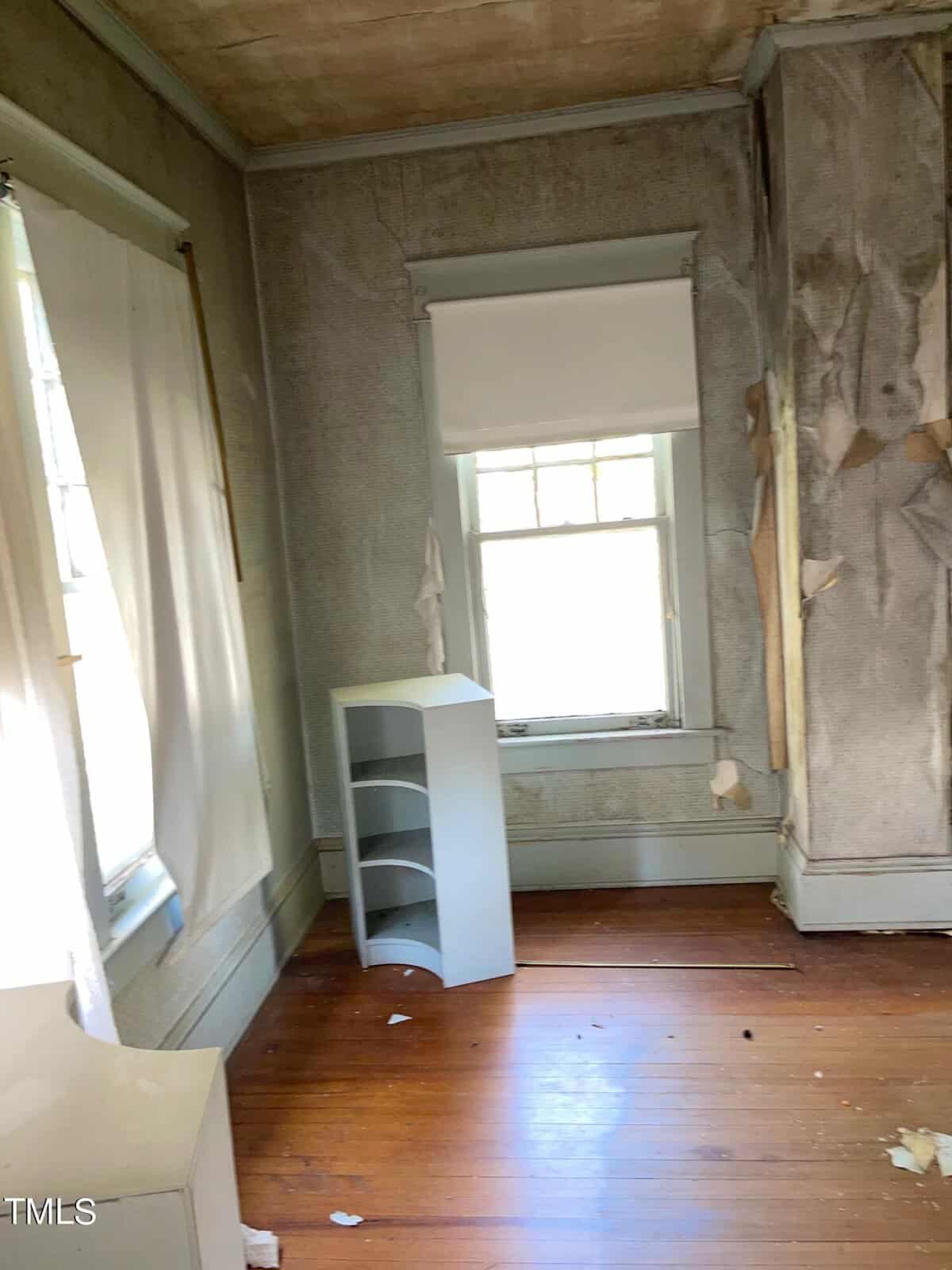 A room with peeling paint on the walls and ceiling. There are two windows with tattered curtains, and a small white shelving unit beside one of the windows. The floor is wooden and appears worn. The room has an overall abandoned or neglected feel.