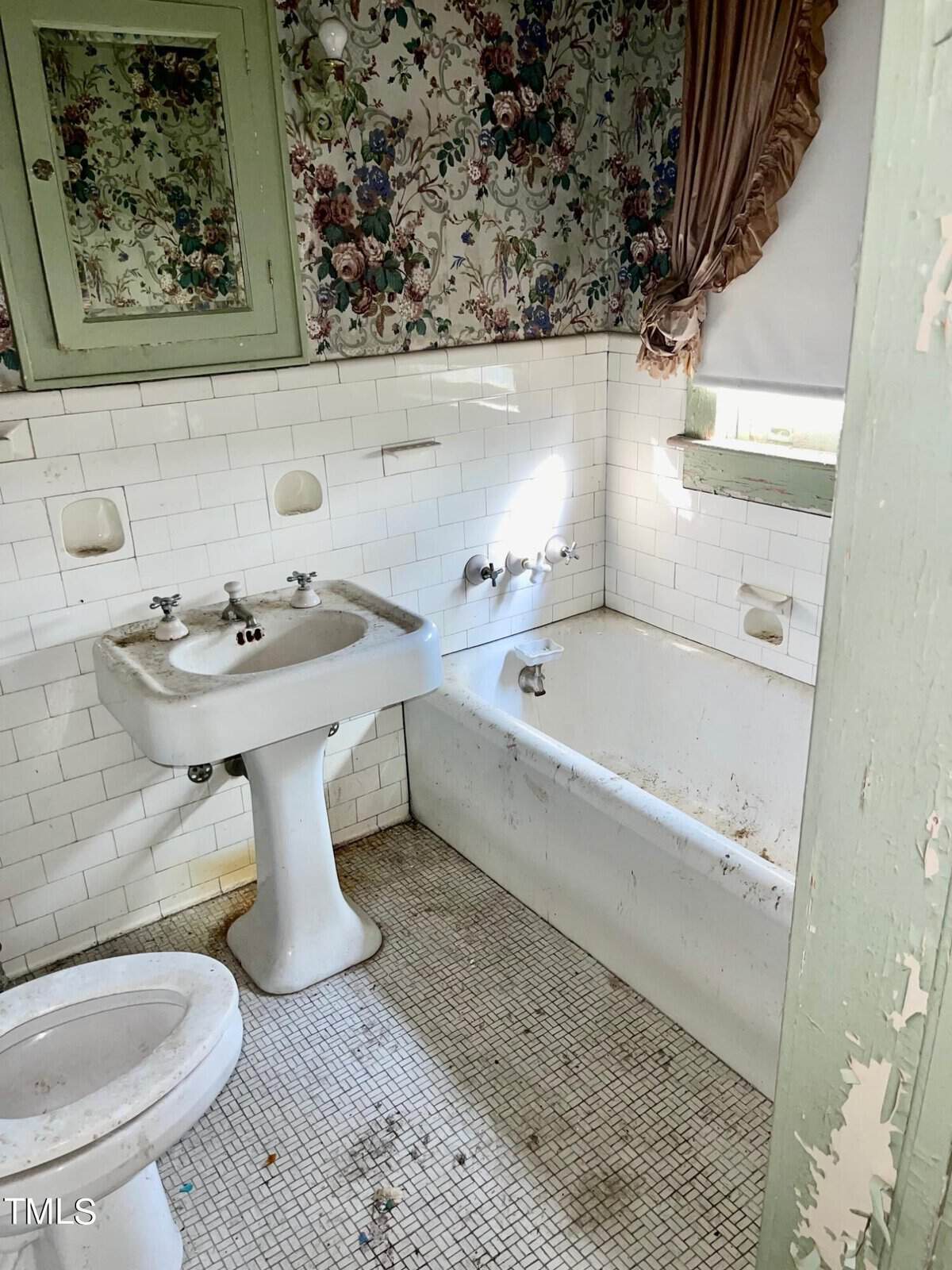 A small, dated bathroom with floral wallpaper, white subway tiles, and a white pedestal sink. The bathtub has visible rust stains and the toilet appears unclean. A window with a brown curtain filters light into the otherwise neglected space.