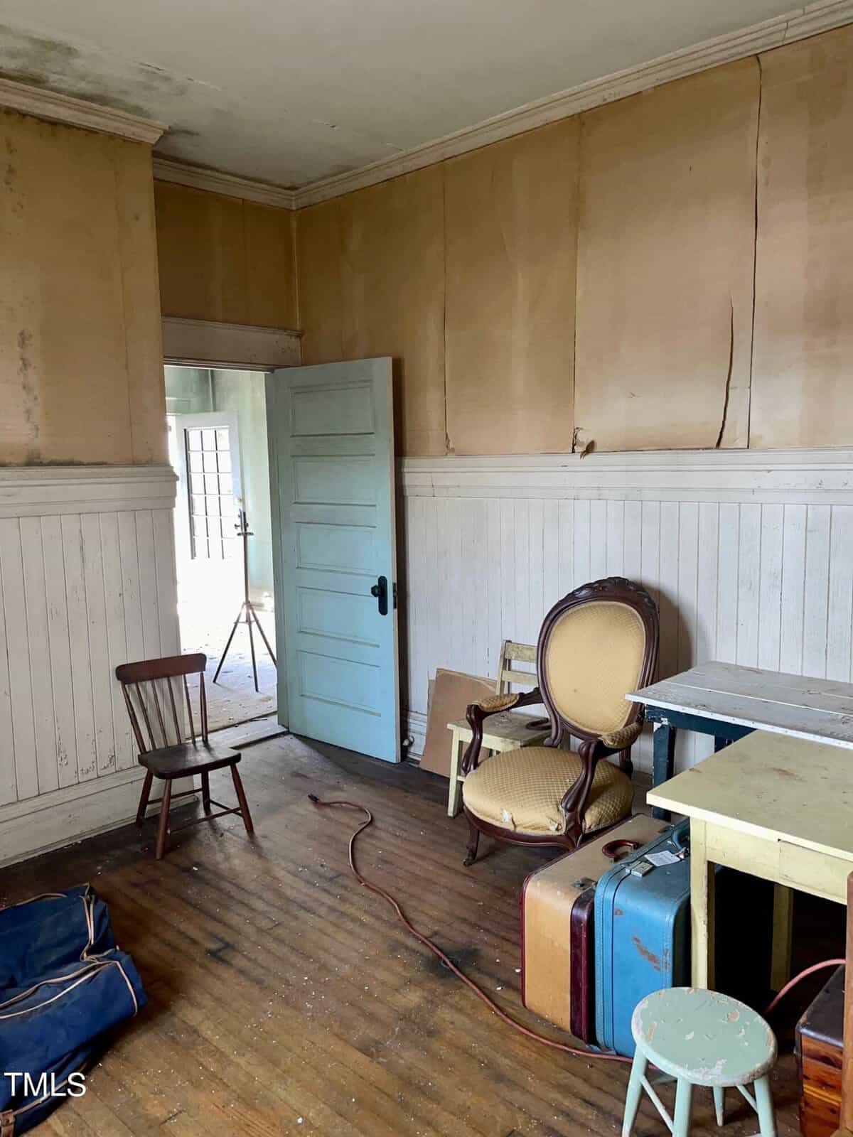 An interior of an old, partially empty room with beige walls and worn wooden flooring. Furniture includes a blue cushioned wooden chair, a round-backed armchair, a small desk, a white stool, and vintage suitcases. A door is slightly ajar, revealing another room.