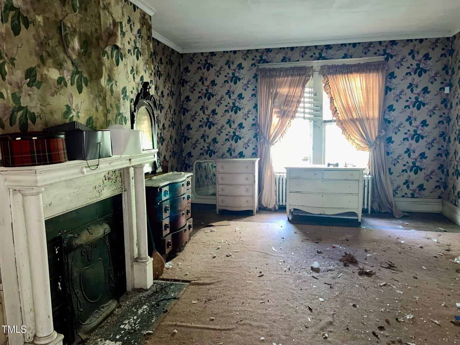 A dilapidated room with peeling floral wallpaper, hardwood floor covered by a worn, beige carpet, and dusty, white wooden furniture. There's debris scattered on the floor. Light filters through sheer curtains covering a single window, casting a dim glow.