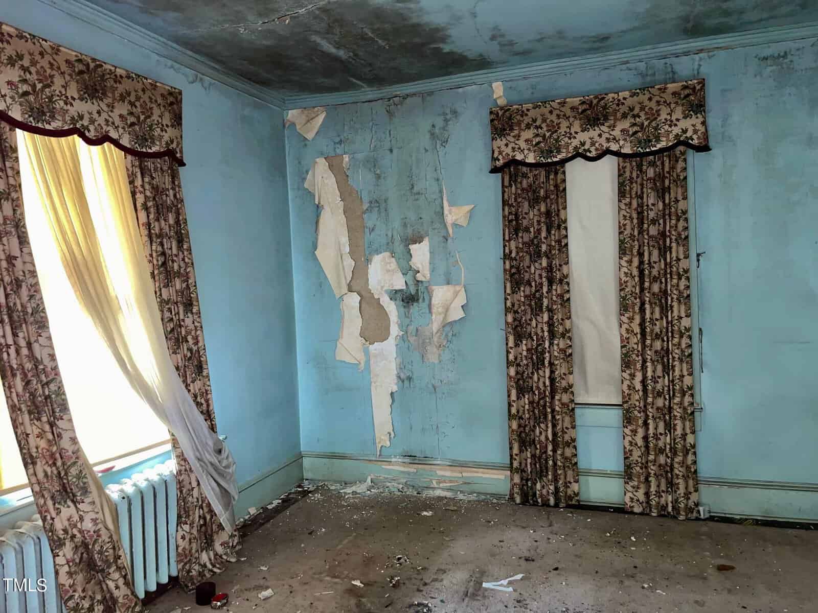 A dilapidated room with light blue peeling walls, stained ceiling, and damaged wallpaper. It has two tall windows with tattered floral curtains. The floor is covered in debris, and there is a white radiator beneath one of the windows.