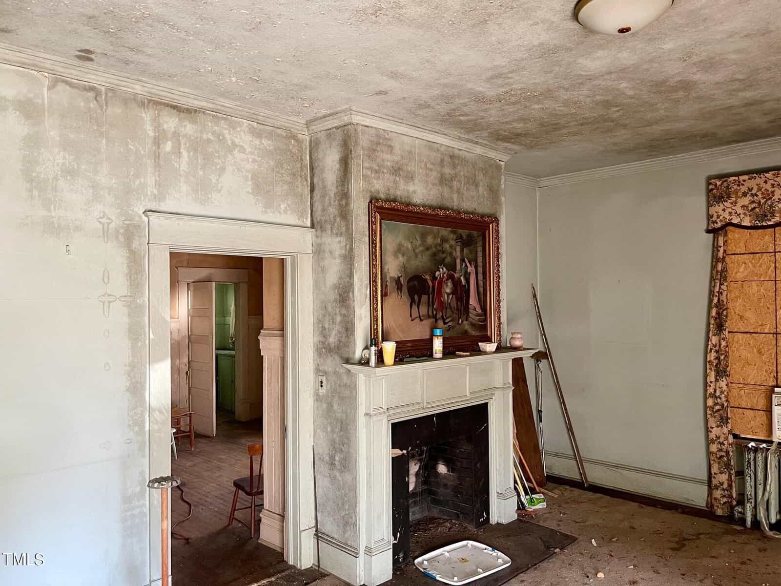 An old, dimly-lit room with a dirty, soot-covered fireplace adorned with a painting above it. The walls are stained, and there are various tools and clutter on the floor. An open doorway leads to another room, where a green cabinet is partially visible.