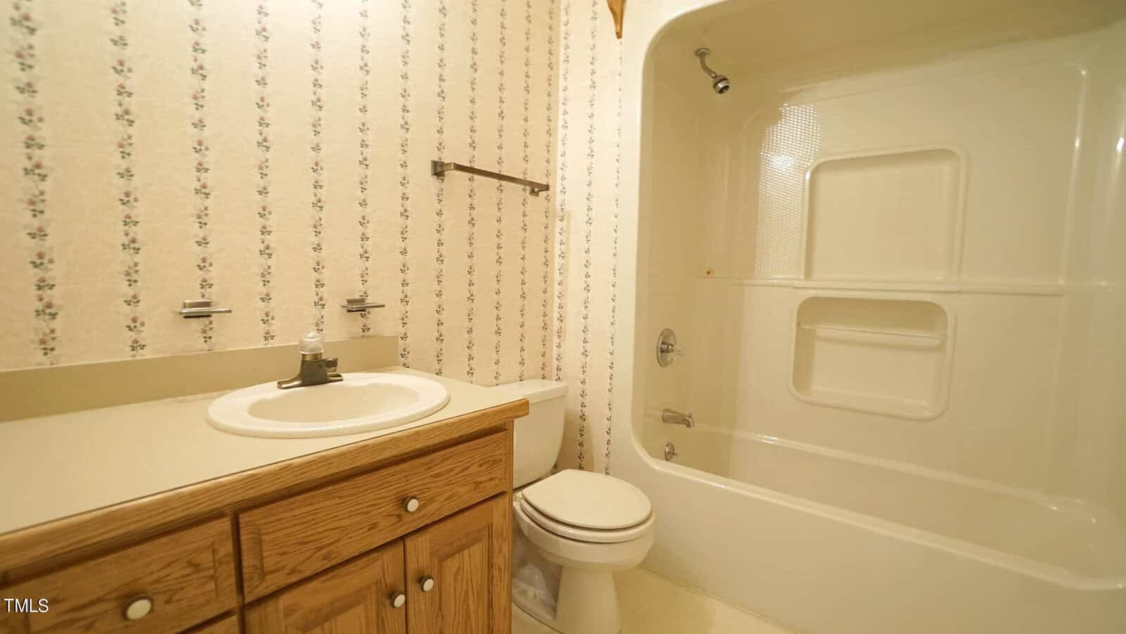 A bathroom featuring floral wallpaper, a wooden vanity with a white sink, and a toilet. The bathtub and shower are combined with built-in shelving. A towel bar is above the toilet.