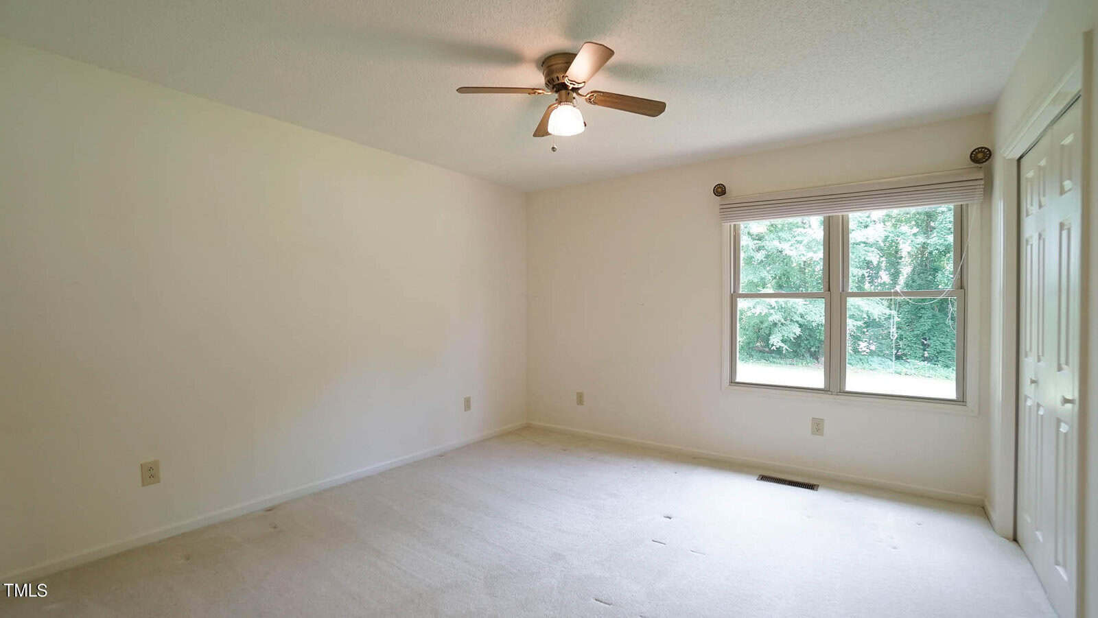 An empty room with light-colored walls and carpeting features a ceiling fan with a light. A large window with closed blinds overlooks a green outdoor space. A closed door is visible on the right wall.