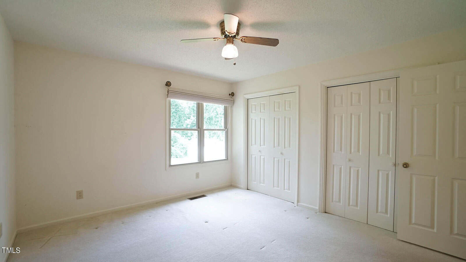 A spacious empty room with cream-colored walls and carpet. A ceiling fan with a light fixture is centered above. There is a large window letting in natural light and two closed closet doors on the right wall.