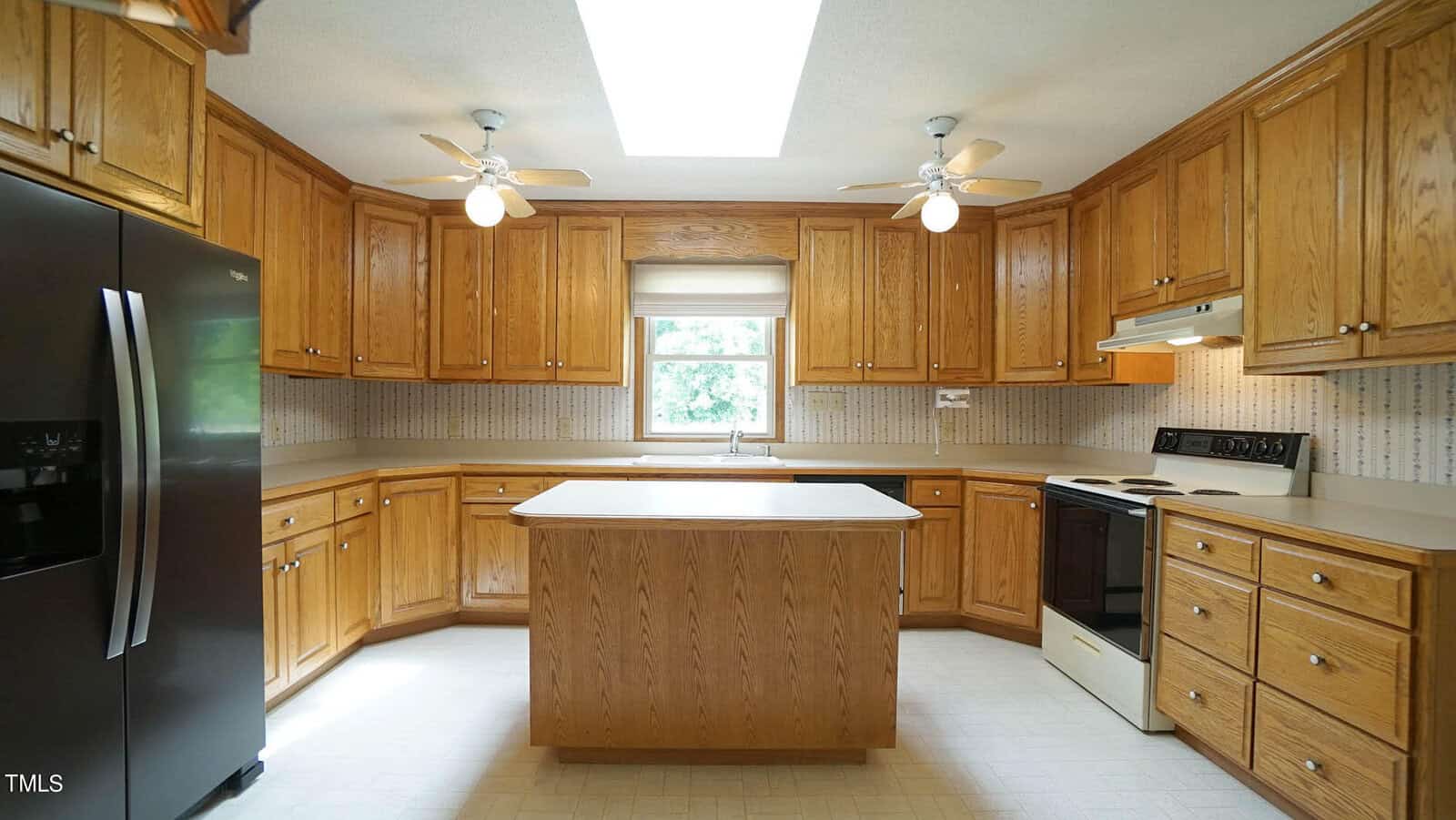 A spacious kitchen with wooden cabinets, a central island, and a white countertop. It features a black refrigerator on the left, and a white stove and oven on the right. Two ceiling fans and a skylight provide lighting. A window is above the sink.