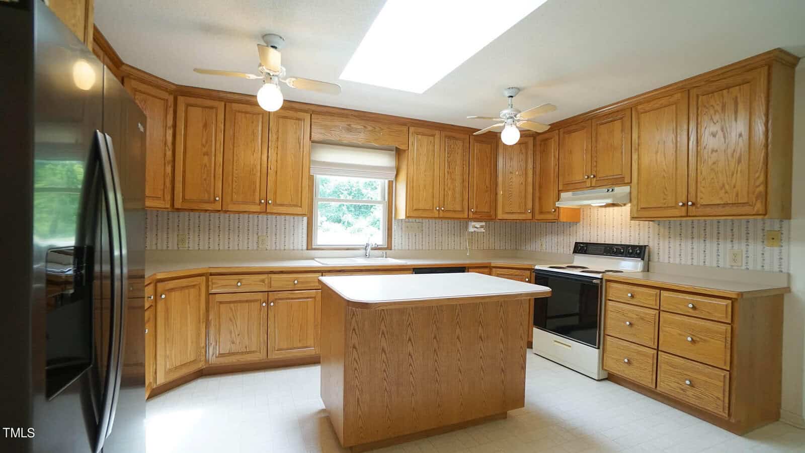 A spacious kitchen with wooden cabinets and a central island. Two ceiling lights with fans illuminate the room. A white stove and a black refrigerator are visible, along with a window offering natural light. The walls have a subtle patterned design.