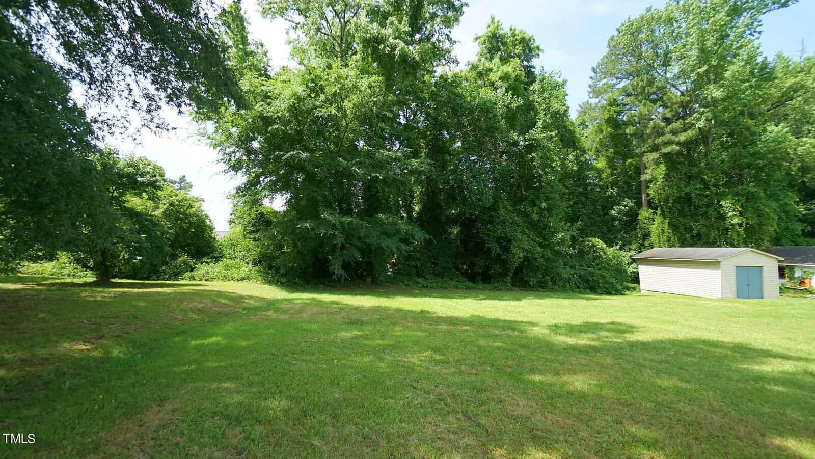 A lush, green yard under a clear blue sky features a spacious grassy area bordered by tall trees and a small, white shed to the right.