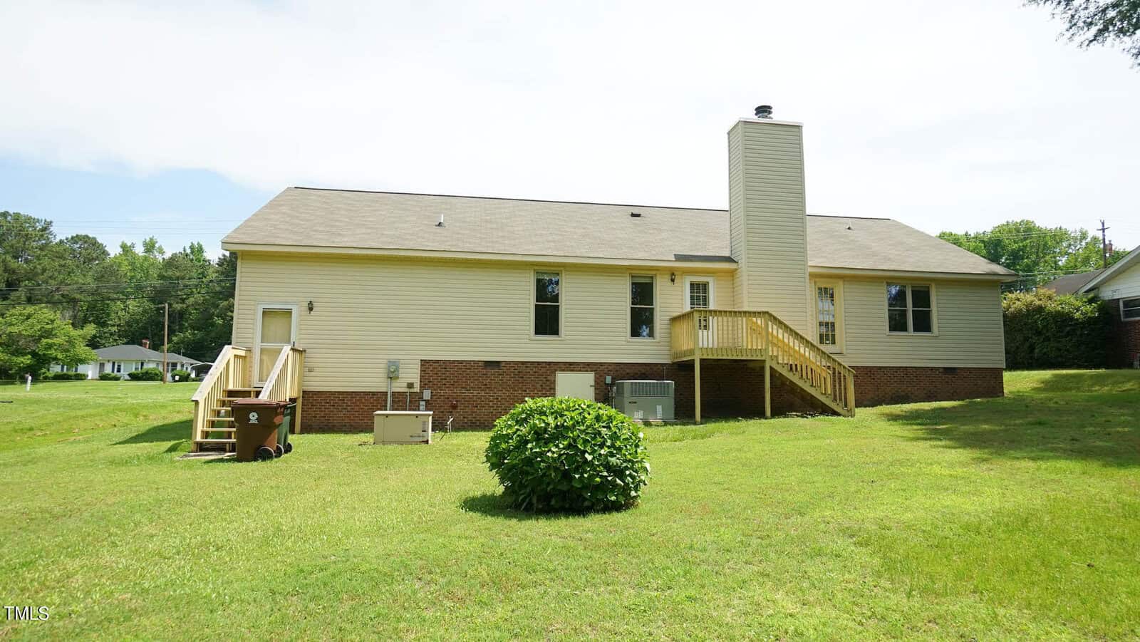 A beige house with a brick foundation, featuring a chimney and two sets of steps leading to an elevated porch. The backyard is grassy with a small shrub in the center. The area is surrounded by trees and a neighboring house is visible in the background.