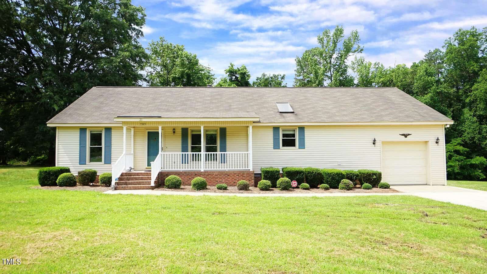 A charming single-story white house on 709 W Lake Road in Creedmoor boasts a front porch, blue shutters, and an attached garage. It's surrounded by a well-maintained lawn, bordered by bushes, with trees visible in the background under a partly cloudy sky.