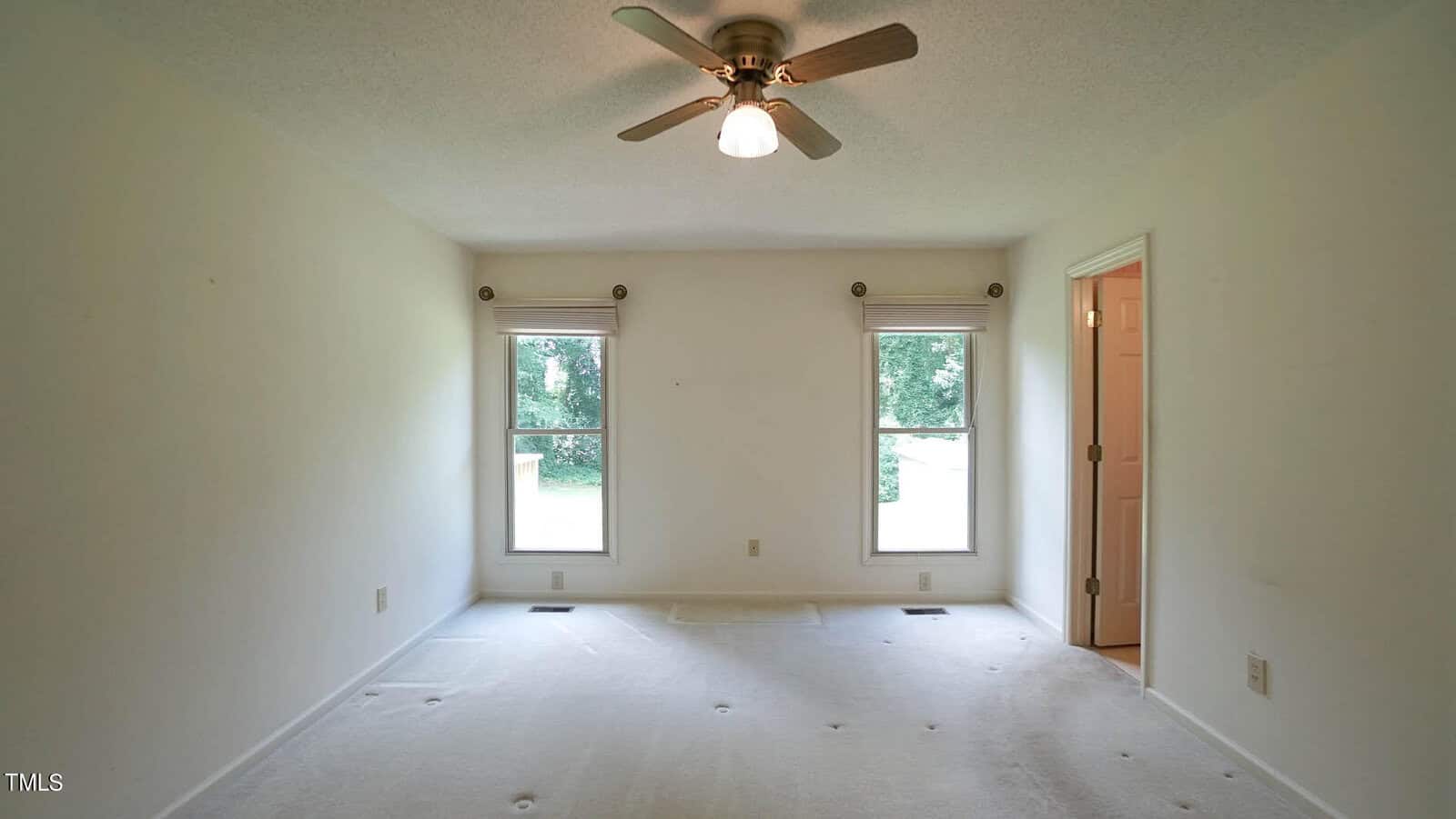 An empty room with white walls and carpet, featuring two windows with roller blinds. A ceiling fan with a light is centered above. A doorway leads to another room. Natural light filters in through the windows.