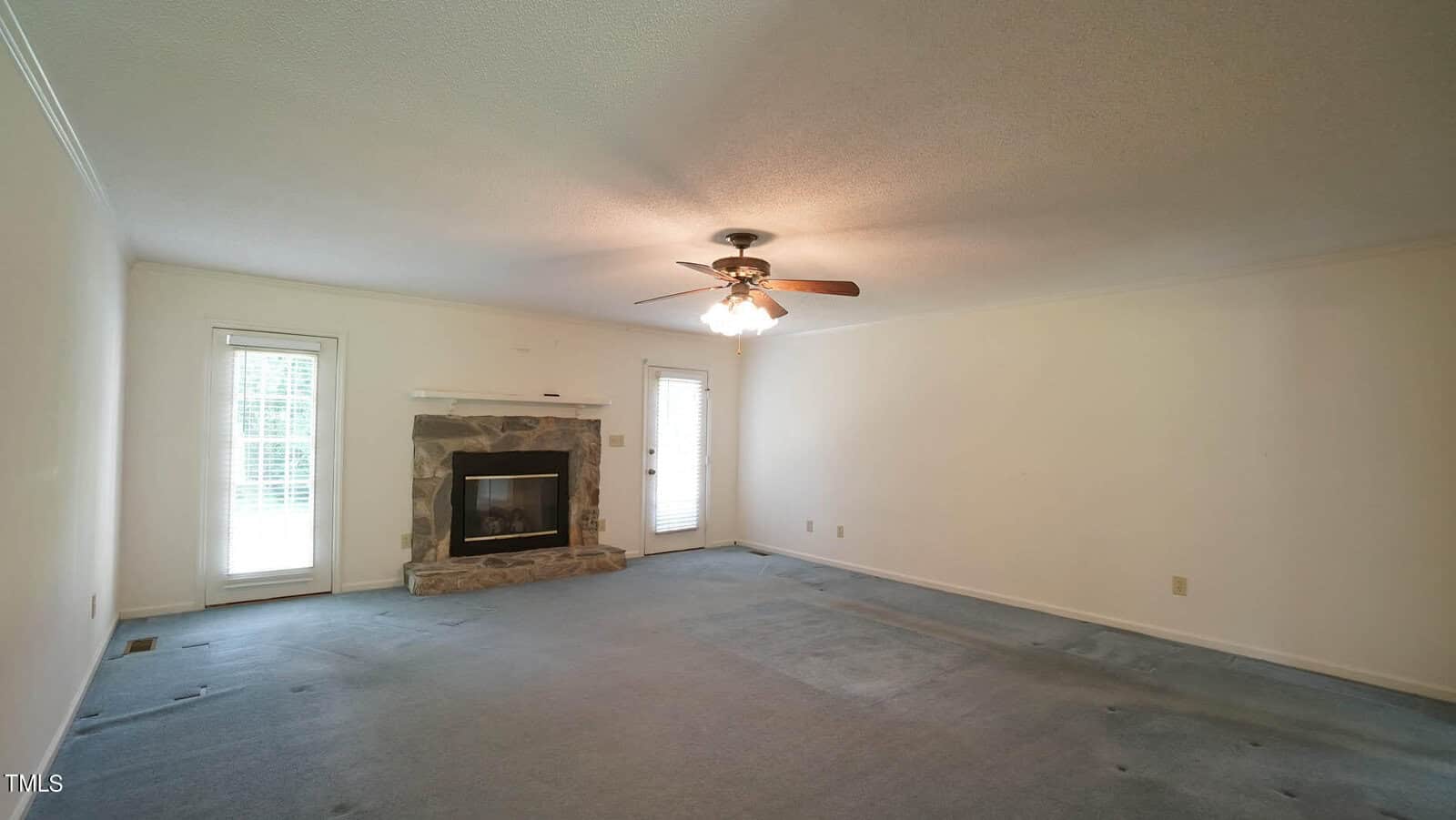 A spacious, empty living room with light beige walls and a textured ceiling. It features a ceiling fan with lights and a stone fireplace. Two tall windows on the left and right allow natural light into the room. The carpet is light blue.