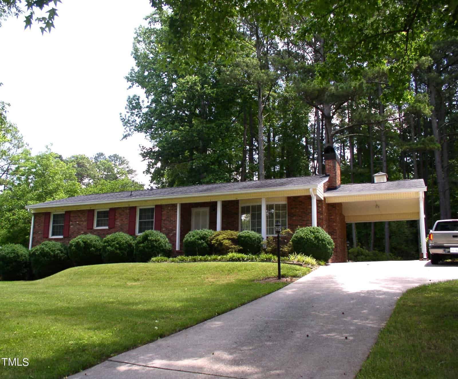 A single-story brick house at 1819 Lynne Avenue in Henderson, NC, boasts a well-manicured lawn surrounded by tall trees. The carport shelters a vehicle, while a concrete driveway leads up to the house. The front yard features a charming mix of shrubs and greenery.