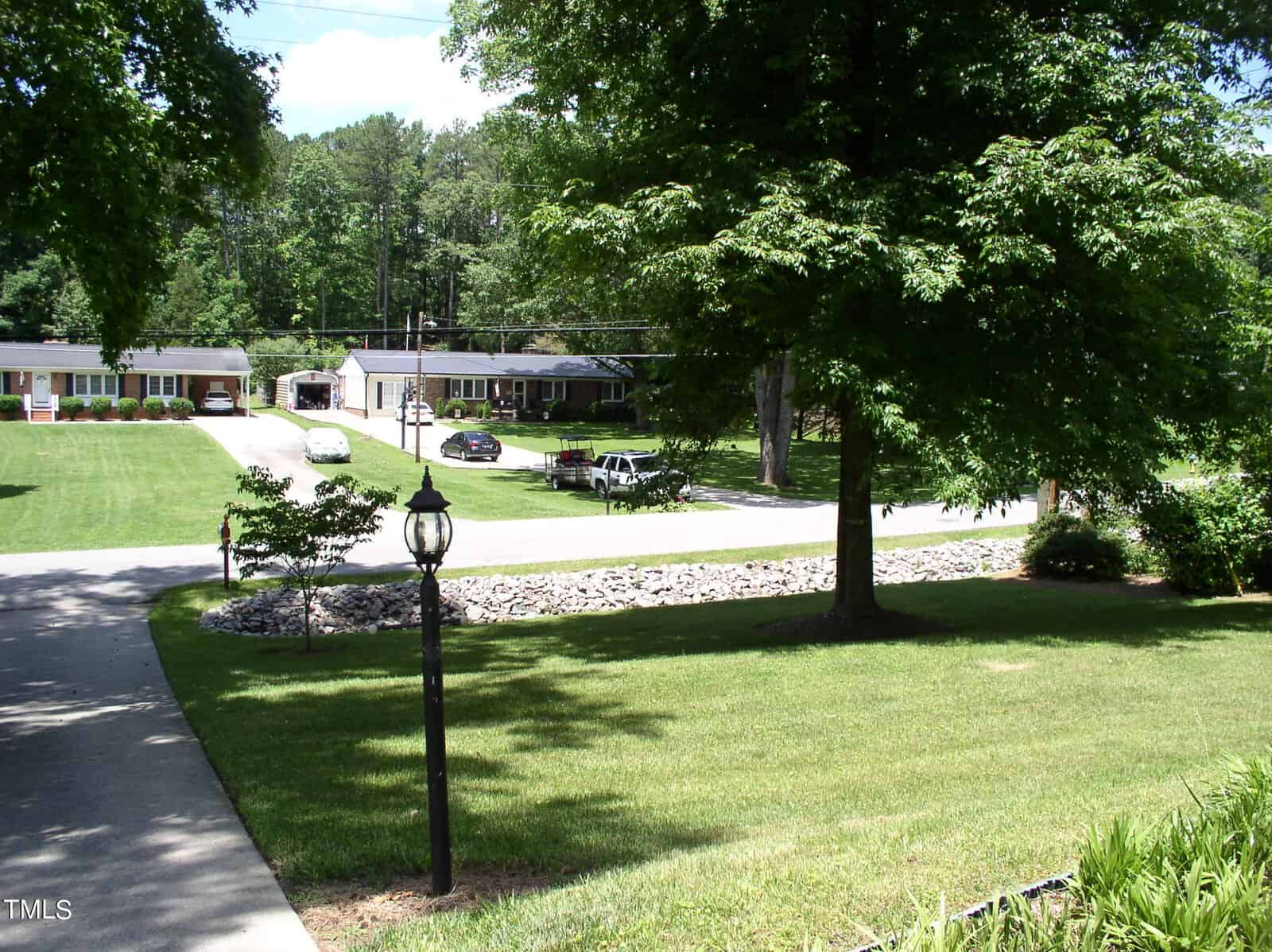 A well-maintained suburban street with green lawns and trees. One side of the street features a row of houses, each with driveways and cars parked outside. A lamppost is visible in the foreground near a paved walkway and road lined with rocks.