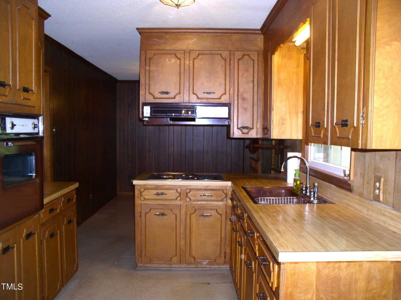 The image shows a vintage kitchen with wooden cabinets and countertops. An electric stovetop is built into the counter, with an oven and range hood above. A double sink with a window above it is situated to the right. The walls and cabinets are a matching wood finish.