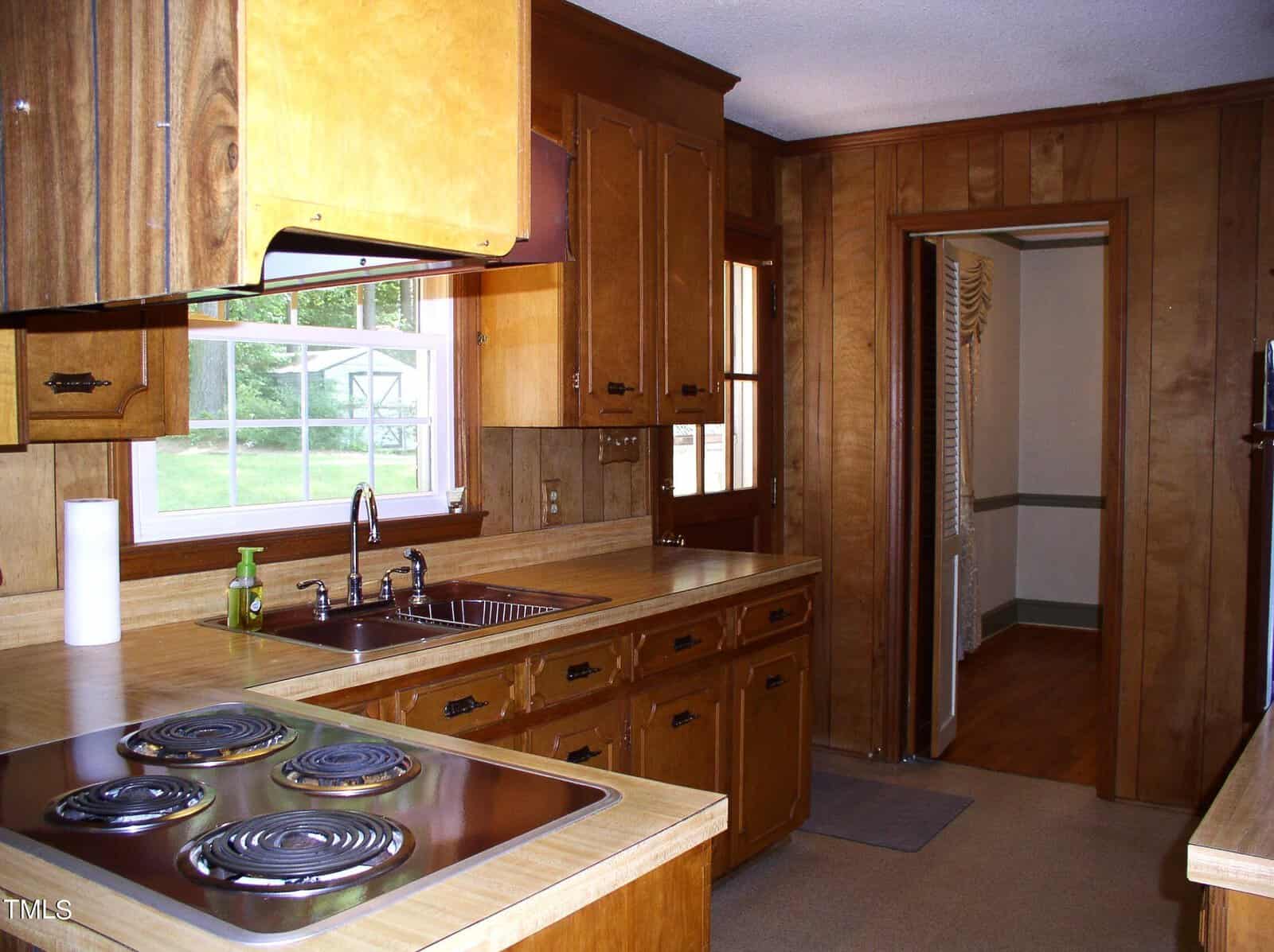A cozy kitchen with wooden cabinets and a beige countertop. On the counter are a paper towel holder, a dish soap bottle, and an electric stovetop with four burners. A window above the sink offers a view outside. The walls and ceiling are wood-paneled.