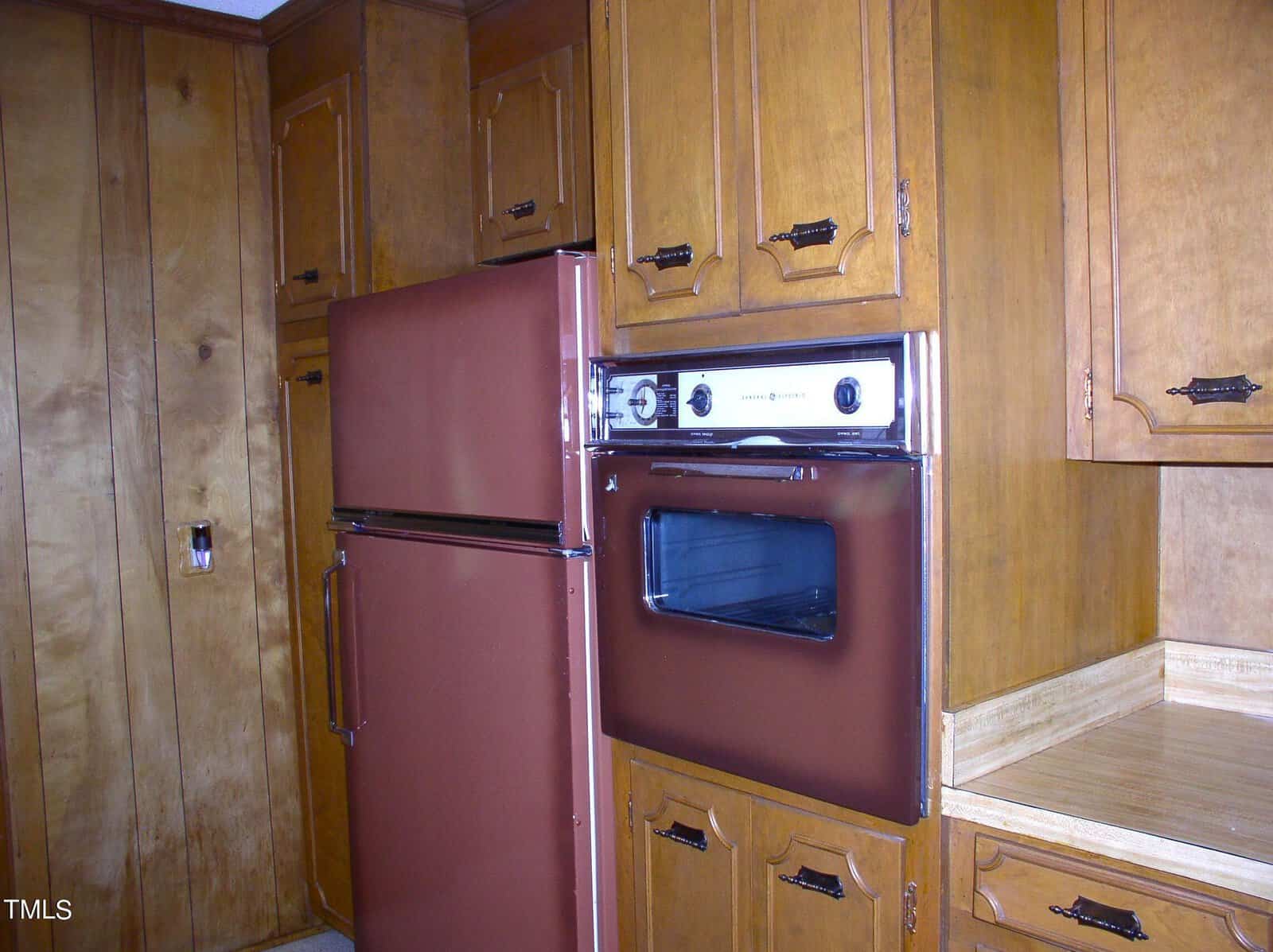 A kitchen with a retro-style design featuring wood-paneled cabinets and burnt orange appliances, including a refrigerator-freezer combo and a wall-mounted oven. The cabinets have ornate metal handles, and there's a small counter space on the right.