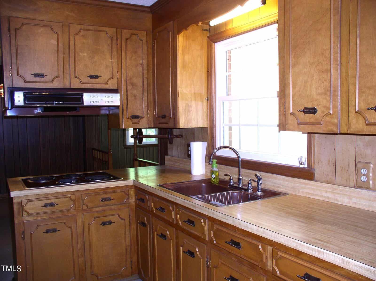 A kitchen with wooden cabinetry and countertops, featuring a stainless steel sink under a window with natural light. There's an electric stovetop embedded in the counter, an overhead range hood, and several drawers and compartments for storage.