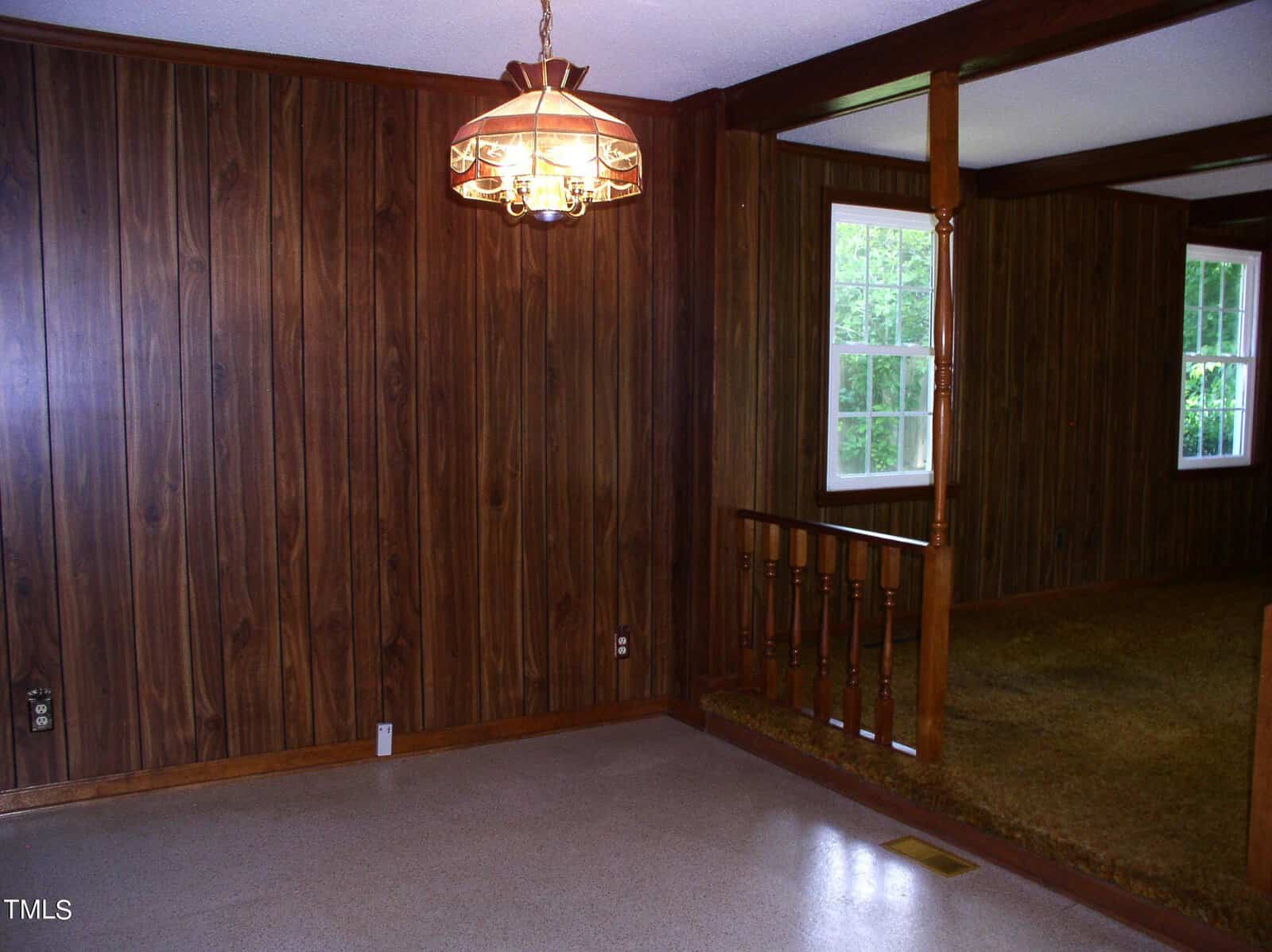 A room with dark wood paneling on the walls and a white ceiling. There is a brass chandelier hanging from the ceiling. A half-wall with wooden railings separates another area with a window that lets in natural light. The floor is mostly bare with a small carpeted area.