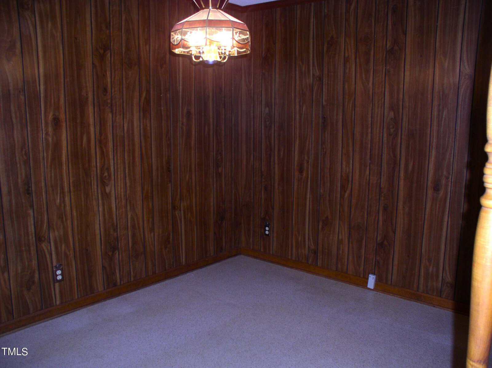 An empty room with dark wood-paneled walls, beige carpeted floor, and a chandelier with a glass and metal design hanging from the ceiling. There are electrical outlets on the walls, with one outlet visible on each of two adjoining walls.