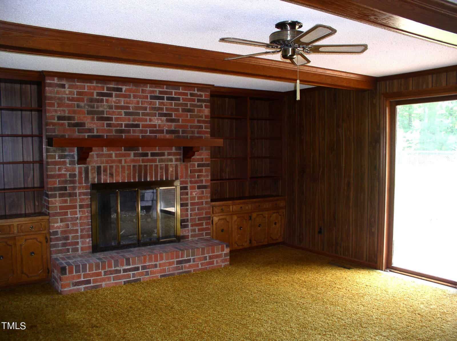 A living room with a brick fireplace at the center, flanked by wooden bookshelves. The room has wood-paneled walls, a ceiling fan, and a large window on the right. The floor is covered with yellow carpet.