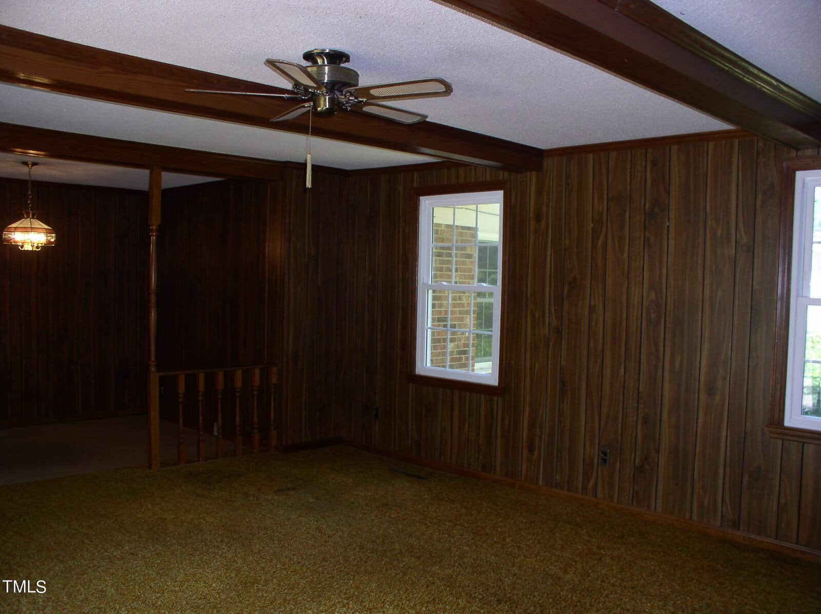 A room with dark wood-paneled walls, a ceiling fan, and yellowish-brown carpet. Two windows allow natural light in. The room has exposed ceiling beams and leads to another space with a visible hanging light fixture.