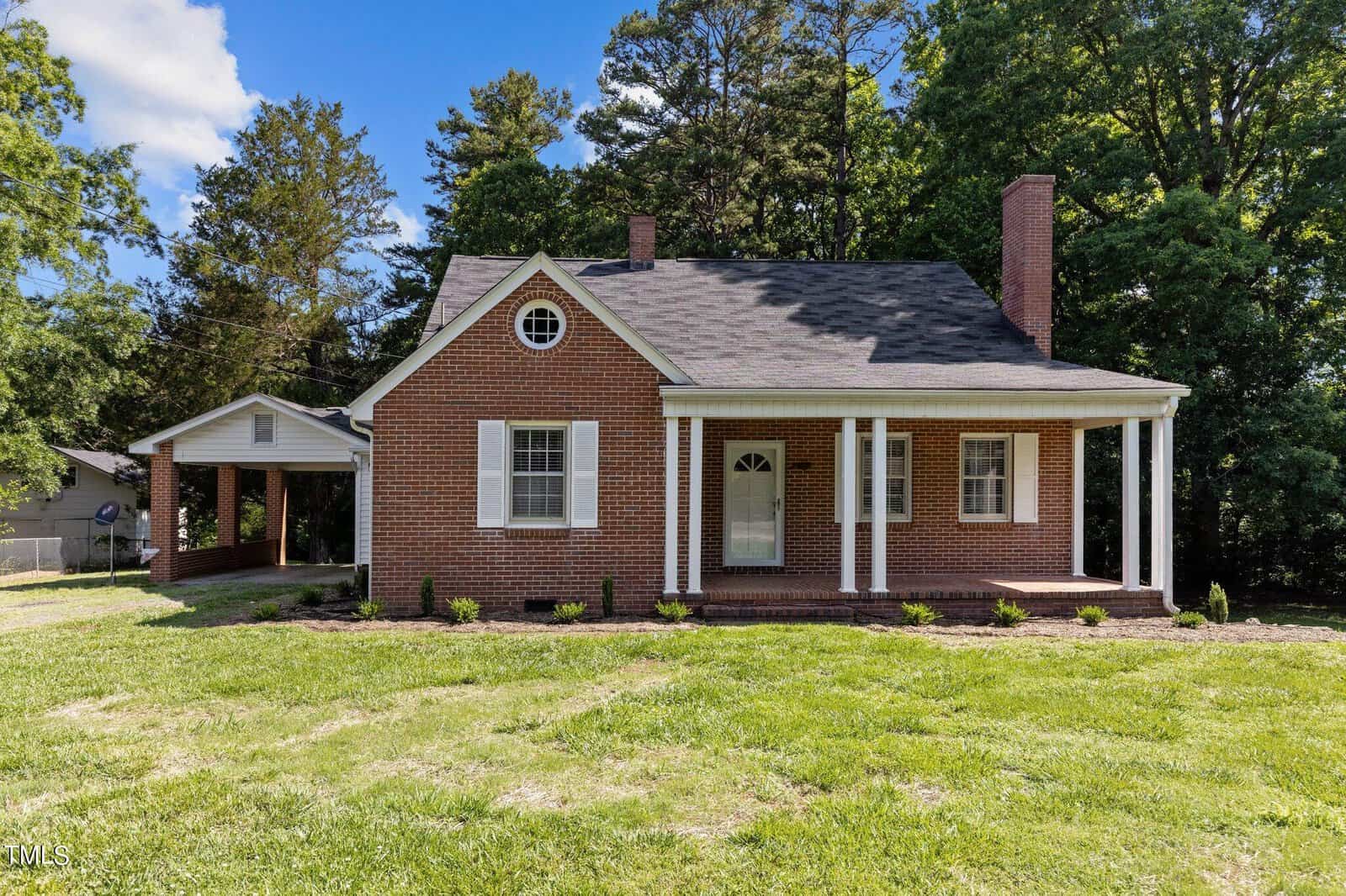 A brick house with a covered porch and two white columns, set against a backdrop of tall trees. It has a gabled roof, white shutters, and a small, manicured lawn in the foreground. A carport is visible on the left side.