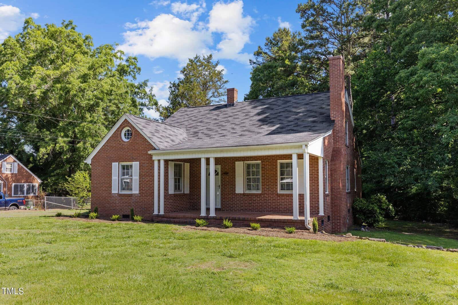 A red brick house with a gabled roof and a white columned porch stands on a grassy lawn. Trees and a partly cloudy sky are in the background. Another house is visible to the left.