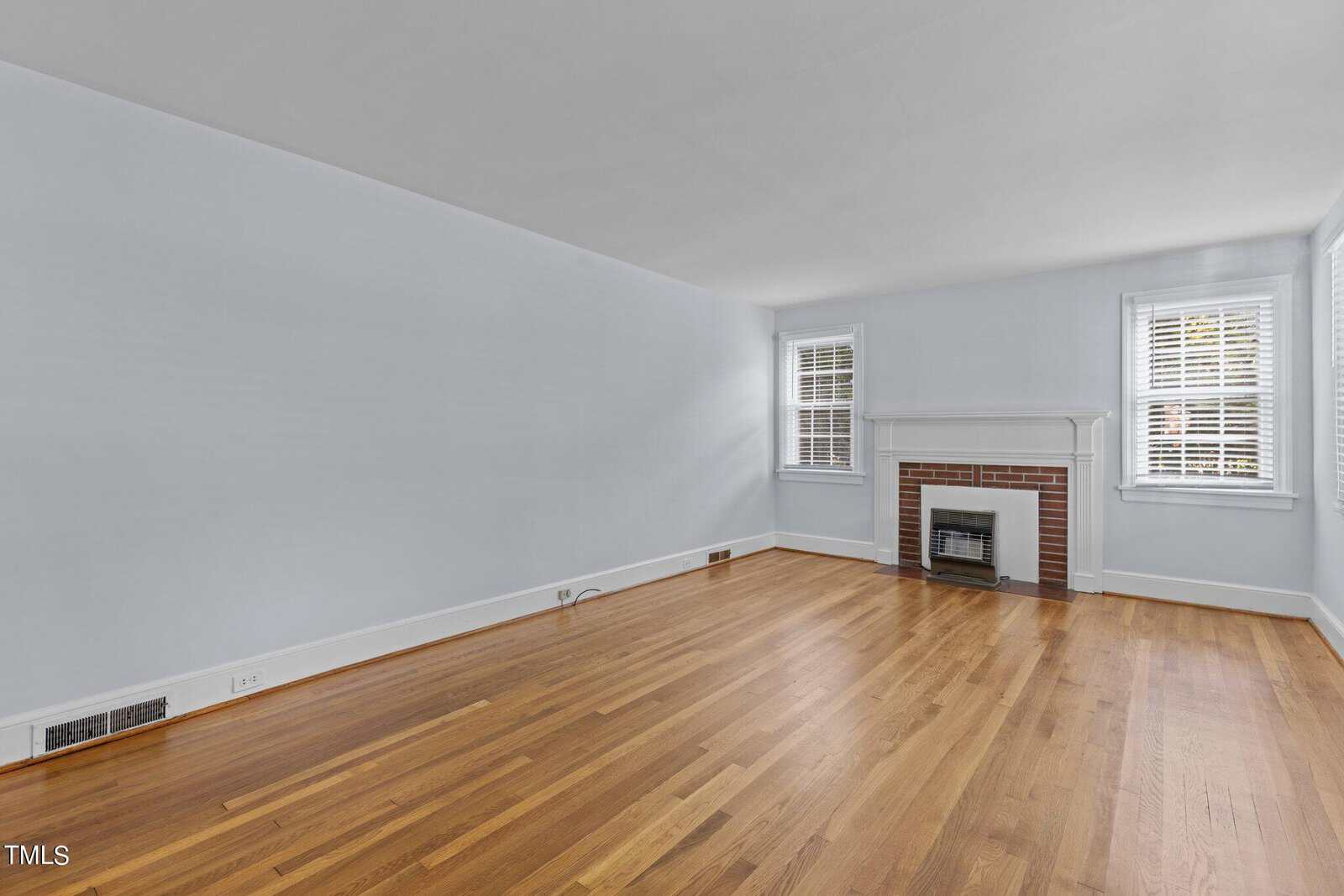 A bright empty room with light blue walls and wooden floors. It features a white-trimmed fireplace with a brick surround. Two windows with white shutters let in natural light.