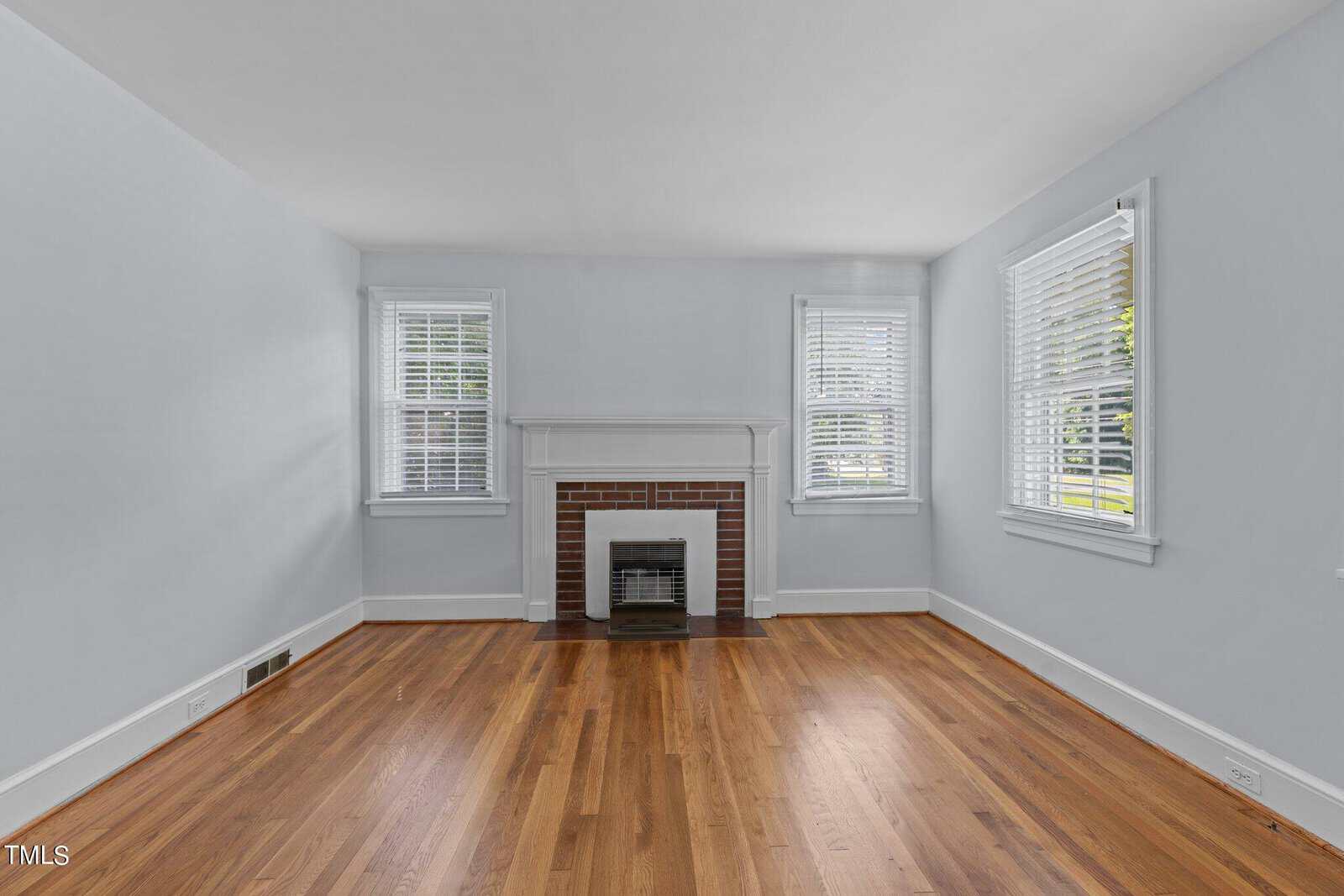 An empty room with light blue walls, hardwood floors, and a brick fireplace centered on one wall. Two windows with white blinds flank the fireplace, letting in natural light. The ceiling is white, and the overall atmosphere is bright and airy.