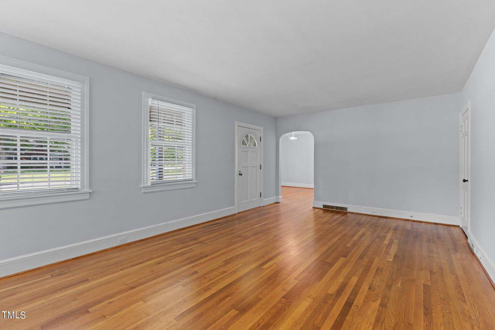 A bright, empty living room with light blue walls, hardwood floors, and two windows with white blinds. A doorway leads to another room with similar flooring. Natural light fills the space, creating a welcoming atmosphere.