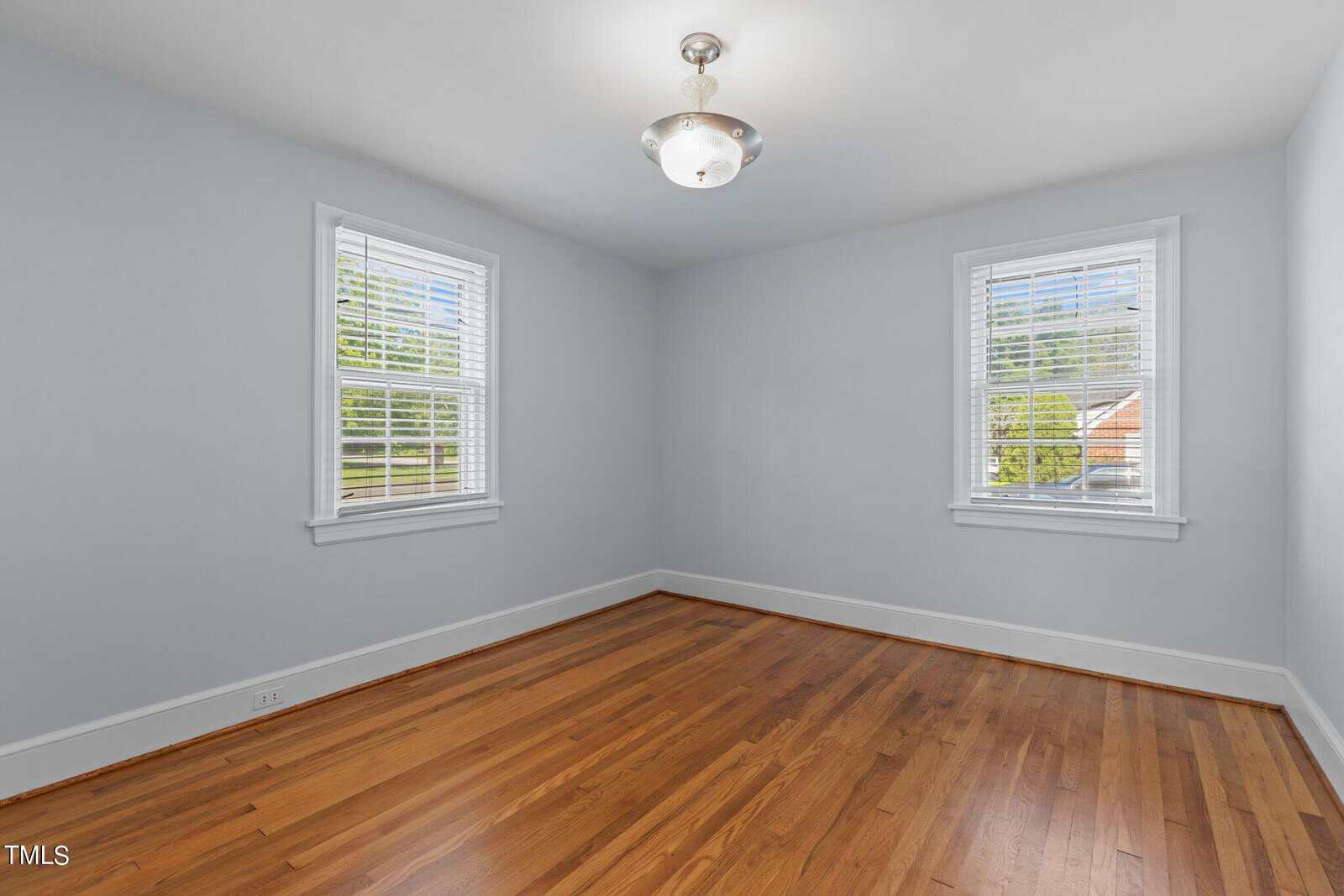 A simple, empty room with light blue walls and hardwood floors. Two windows with white blinds allow natural light to enter. A ceiling light fixture is centered above.