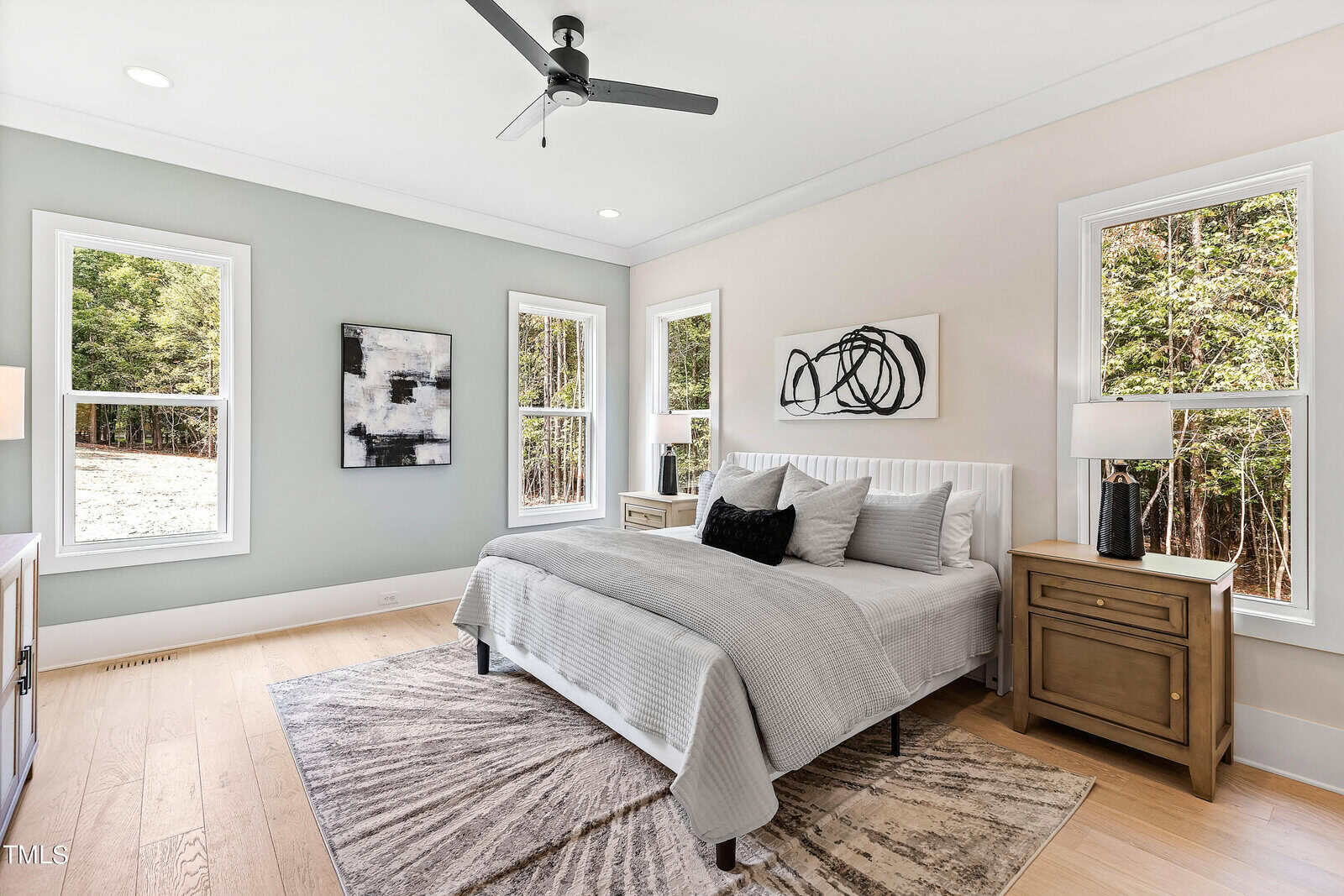 Bright bedroom with large windows, a ceiling fan, neutral bedding, two wooden nightstands with lamps, abstract art on the walls, light wood flooring, and a patterned rug. Trees are visible outside.
