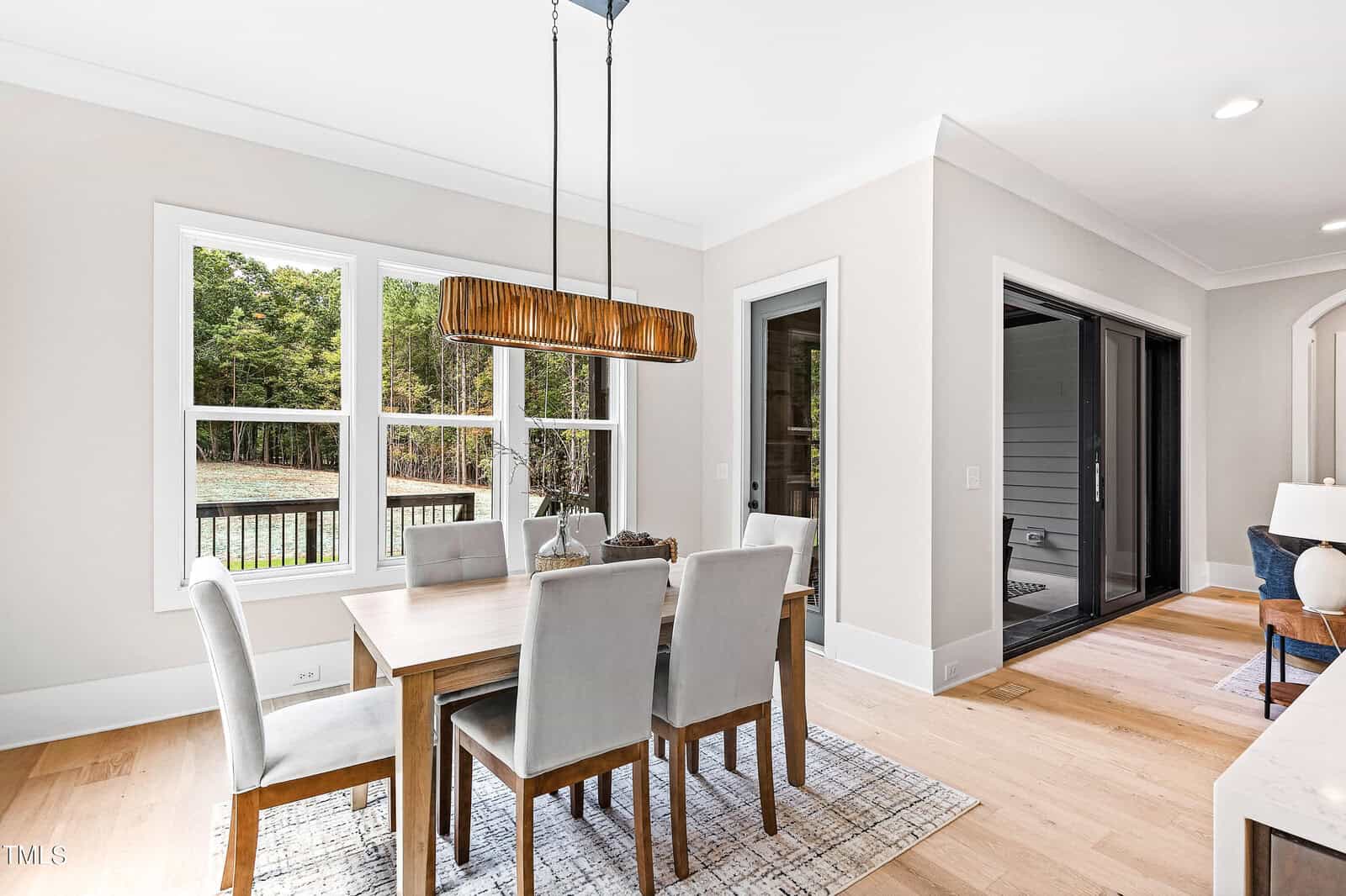 A bright, modern dining room with a wooden table, six upholstered chairs, a rectangular pendant light, large windows showing trees outside, and sliding glass doors leading to a screened porch.